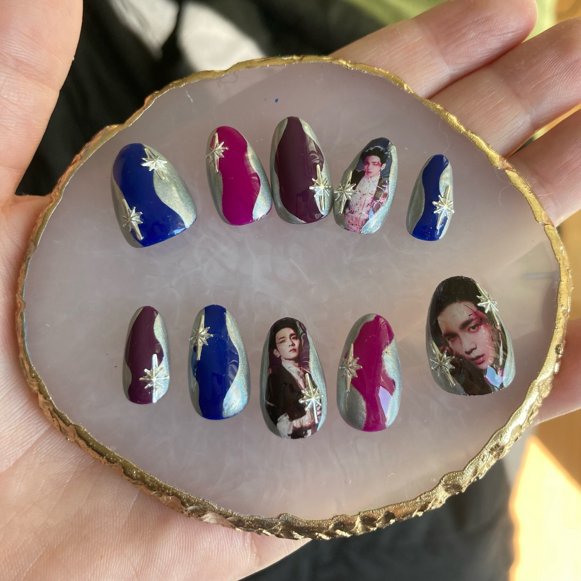 A set of press on nails held on a small resin circle. They are dark toned in the center with silver edges and silver stars. Three of them have images of singer Key from the group Shinee on them