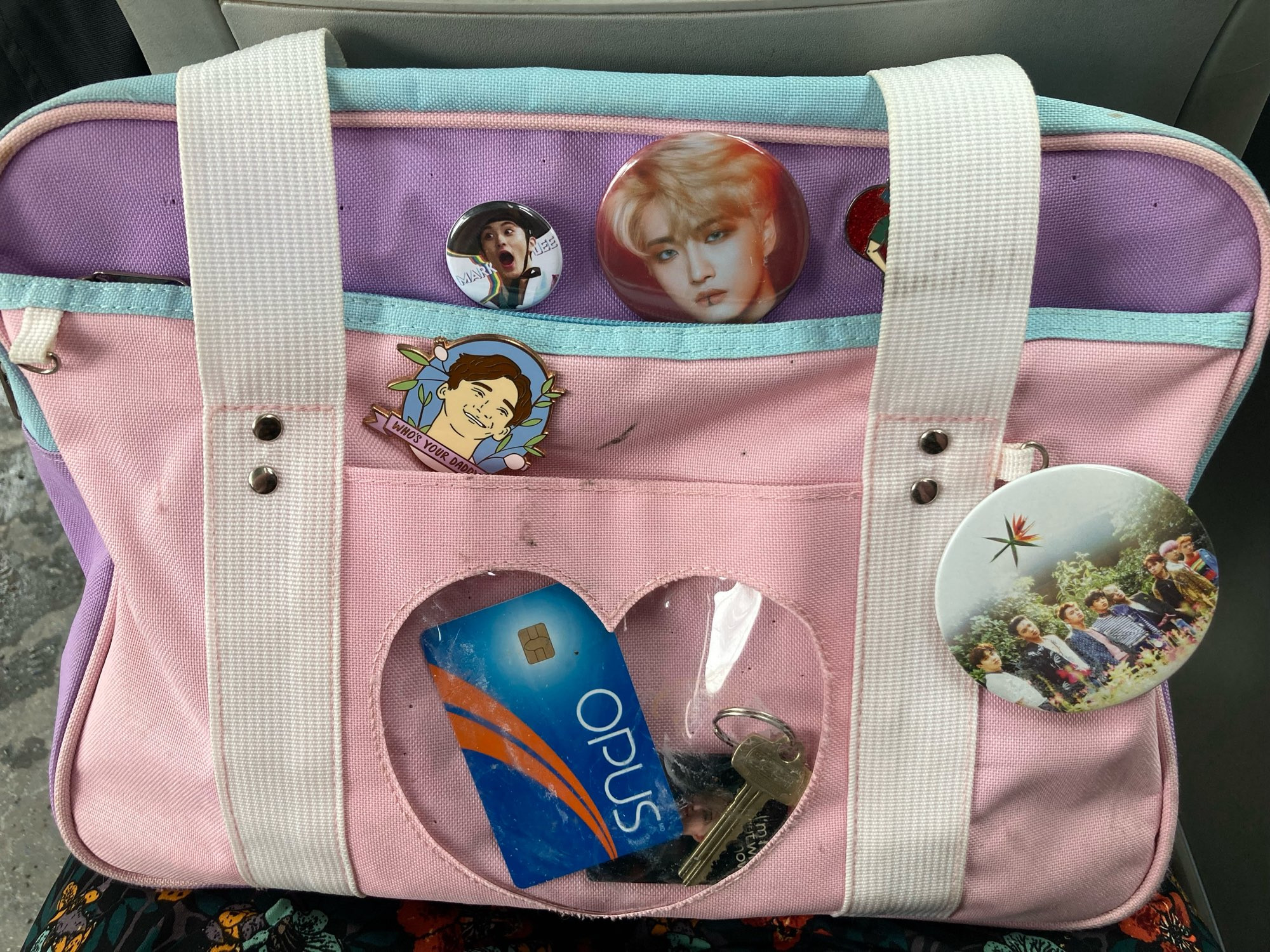 A pink and purple rectangular bag with a heart shaped pocket on the front showing a bus pass and some keys. There are some kpop pins stuck to it and a few grey stains from dropping it in dirty city snow