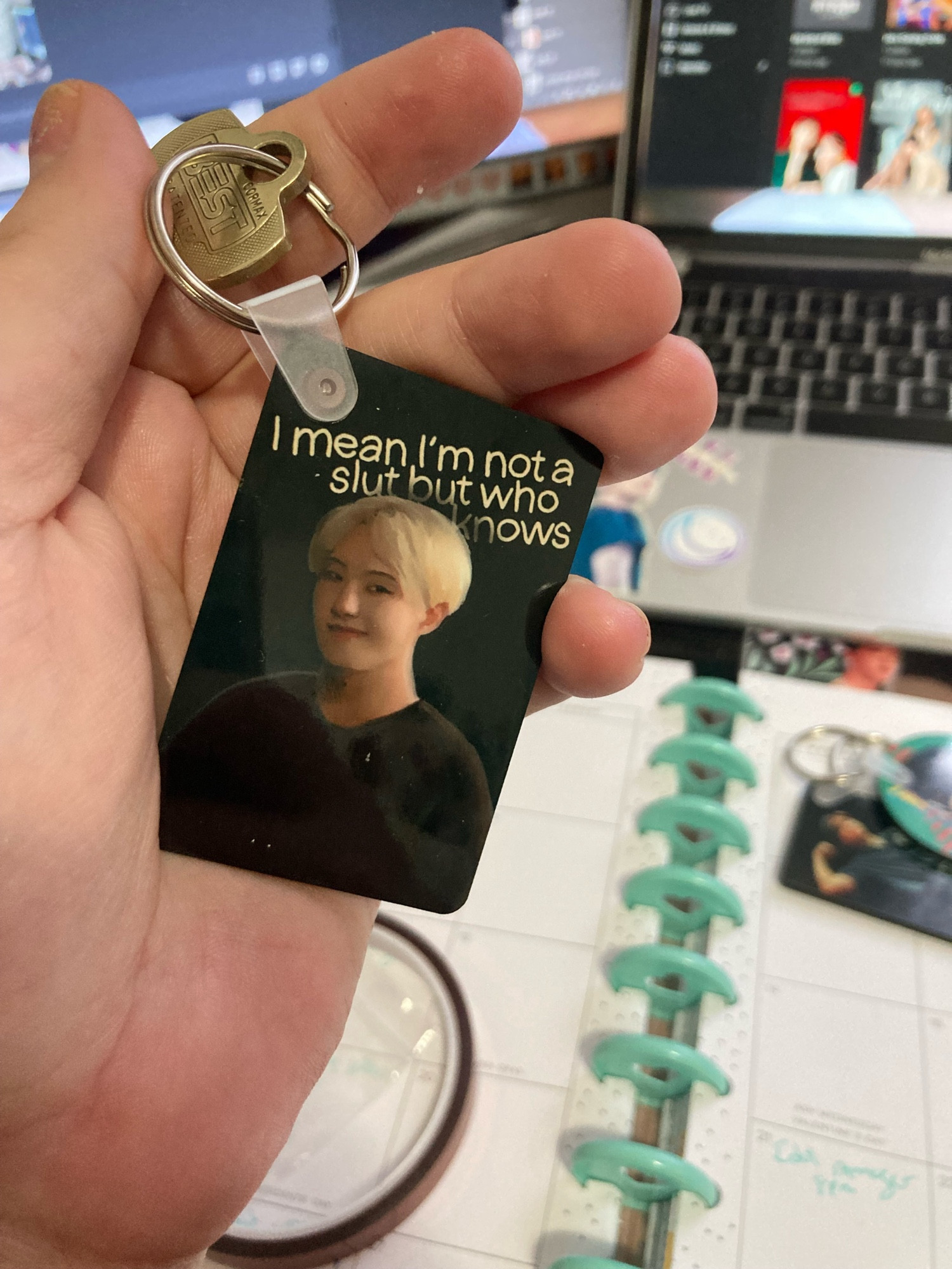 A hand holding a keychain with a painting of singer Hoshi, with ear length blonde hair and a dark sweatshirt with the words “i mean im not a slut but who knows” written behind him