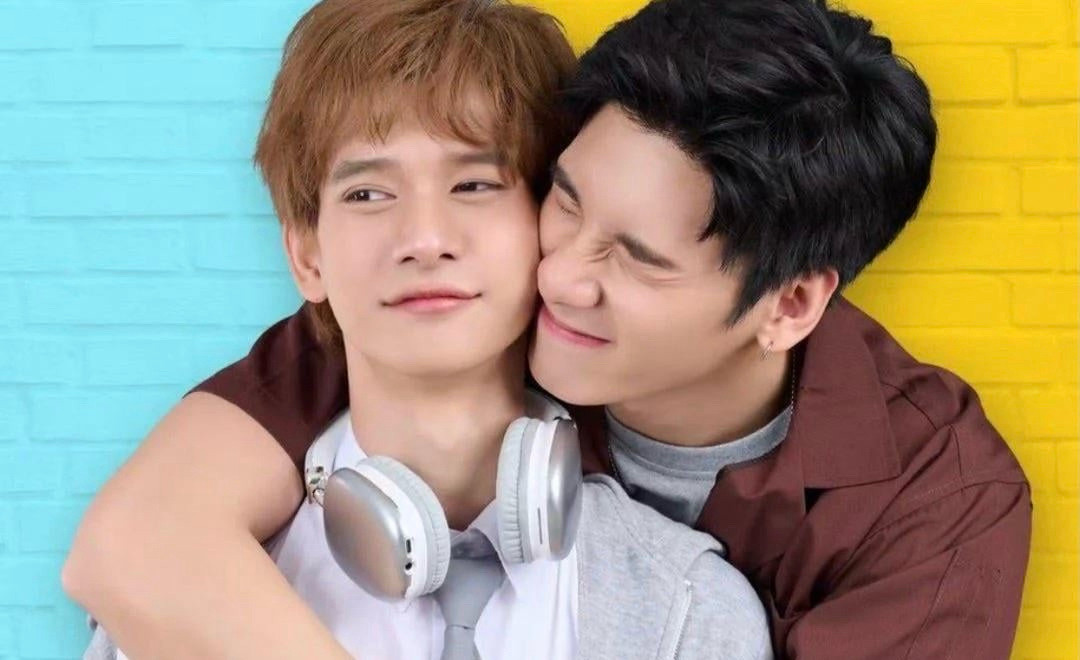 A young man with light brown hair and a pair of headphones around his neck looks off to the side as a dark haired man is hugging him from behind, squishing their faces together affectionately. They are on a background that is half bright turquoise and half bright yellow.