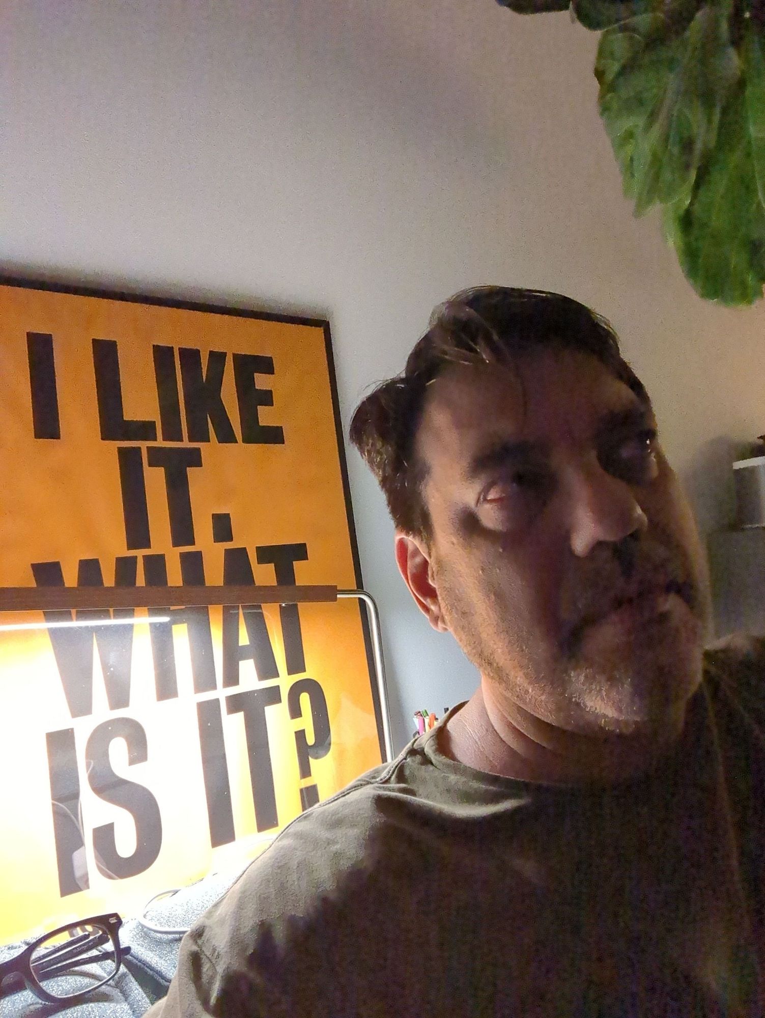 Mid-age man looking up and away from the camera. Pronounced under-eye pockets which run in the family compounded by the late hour. Anthony Burrill's all type/all caps/sans serif I LIKE IT WHAT IS IT? poster illuminated by a bar-shaped desk lamp behind him.