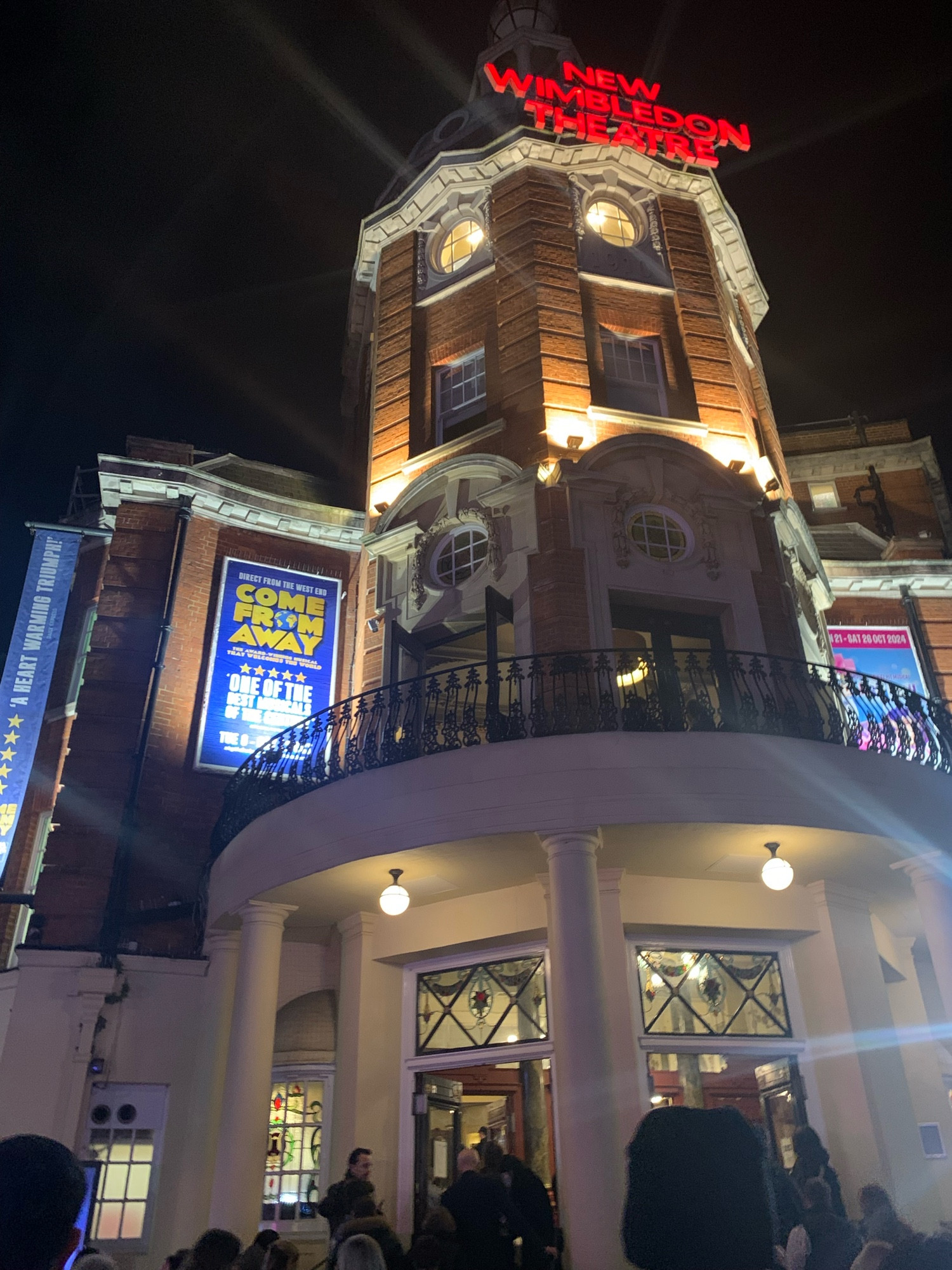 The outside of the New Wimbledon theatre at night. An old (I’m guessing Victorian?) building with its name in red probably-not-actually-neon lights. The audience are walking up stairs and past pillars to a semicircle of doors. There’s a decorative iron balcony above the doors and you can just see the blue and yellow poster for Come From Away to the left of the balcony. 