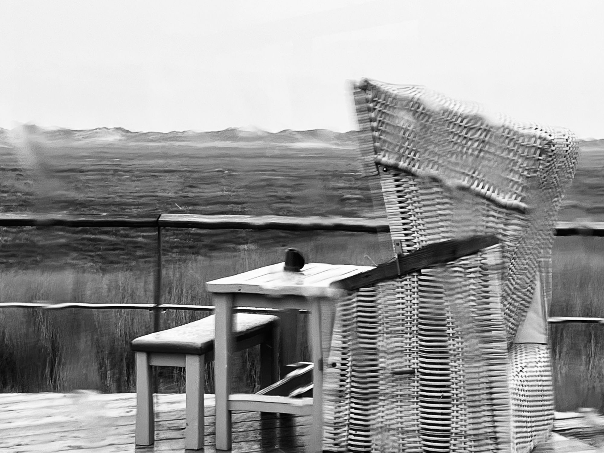 S/W-Bild. Blick durch eine verregnete Glasscheibe auf eine Terrasse, auf der ein leerer Strandkorb mit Café-Tisch steht. Im Hintergrund eine Dünenlandschaft. Durch das an der Scheibe herablaufende Wasser ist die Darstellung verzerrt.