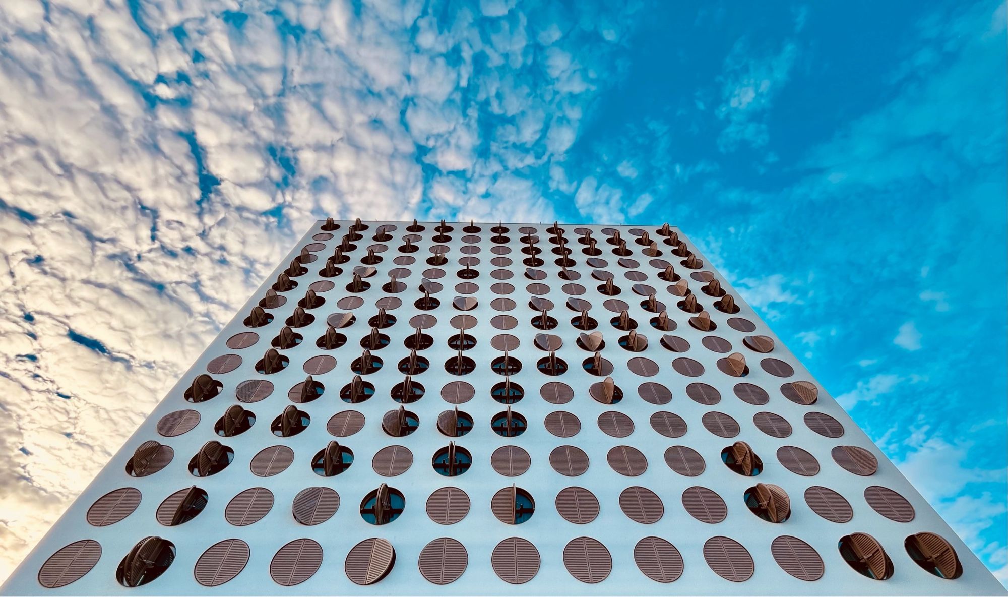 Weitwinkliger Bick Richtung Himmel entlang einer Fassade mit vielen gleichen kreisrunden Fenstern. Einige Fensterläden sind geöffnet. Am Himmel Schäfchenwolken.