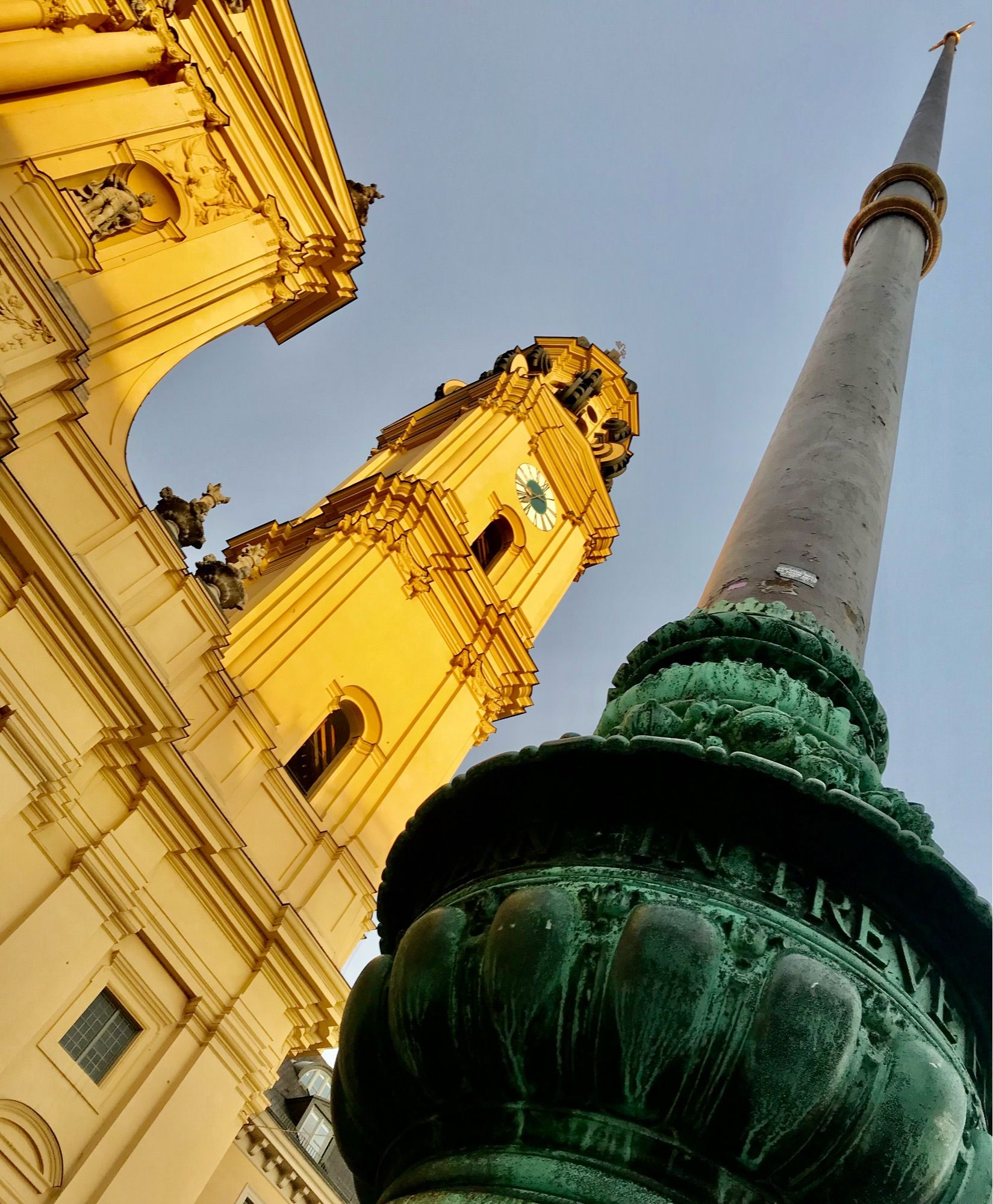 Blick von unten entlang eines Fahnenmasts, der am Sockel reich verziert ist. Links im Bild der Ausschnitt einer ocker-gelb-farbenen Kirchenfront mit einem Kirchturm im warmen Licht der Morgensonne, wolkenloser Himmel.