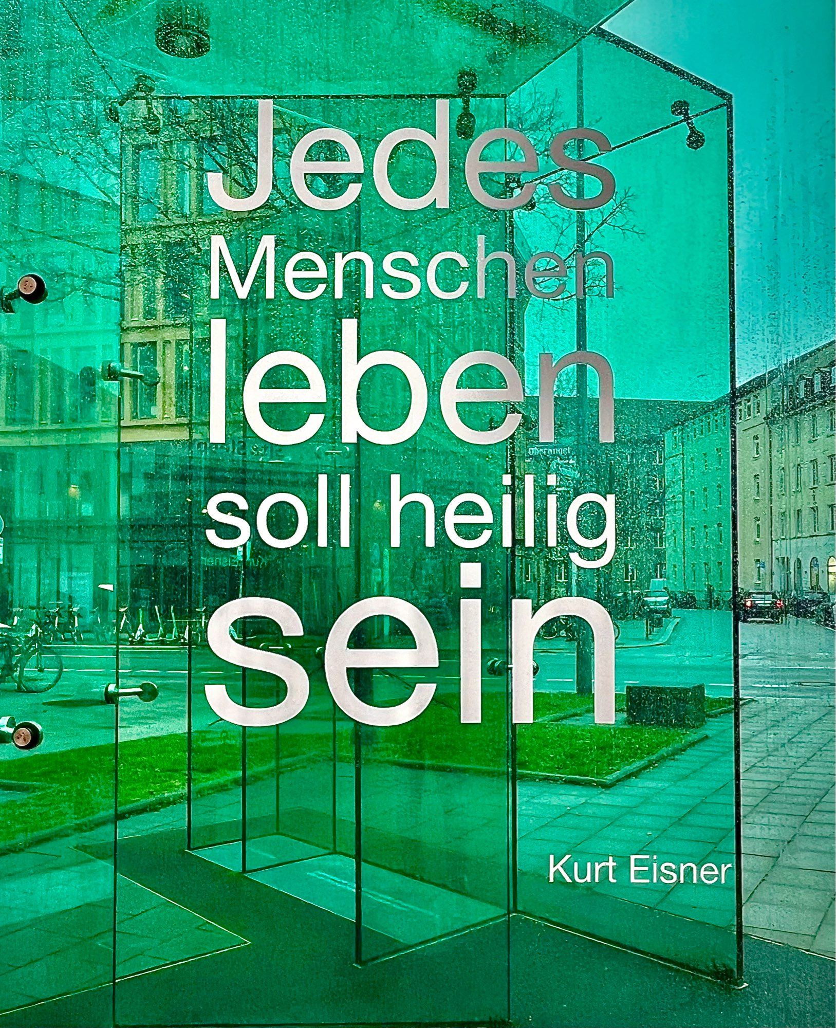 Eine Denkmal aus grünen, transparenten Glasscheiben mit einem Zitat von Kurt Eisner: 
„Jedes Menschenleben soll heilig sein“. Im Hintergrund ist durch das Glas hindurch ein Platz und ein Straßenzug mit Häuserzeilen durch die Glasscheiben erkennbar.