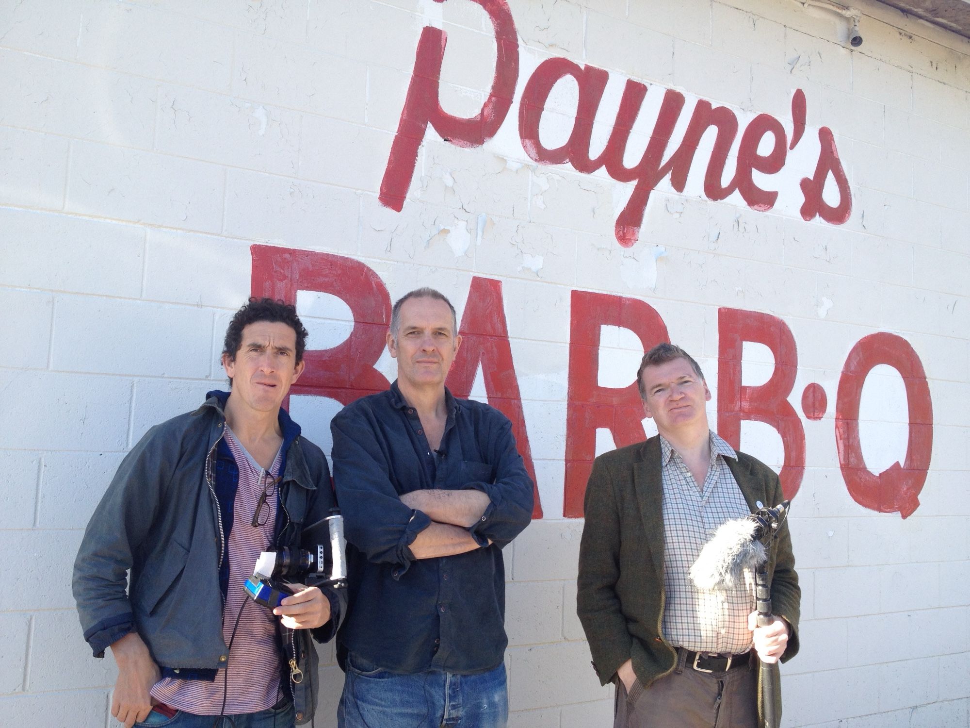 Three middle aged men stand in front of a white wall on which is written in red PAYNE’S BAR B Q