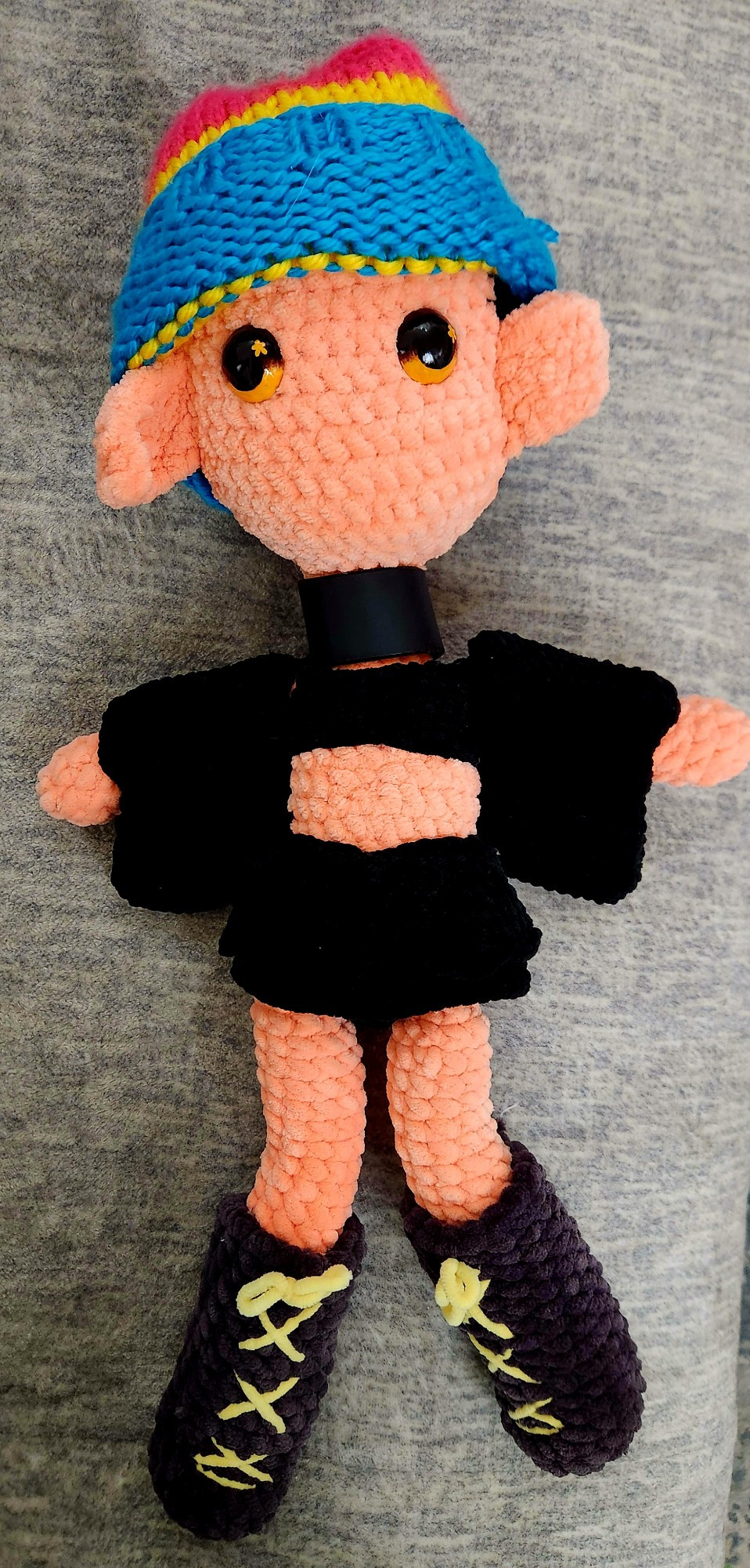 Pictured is a crochet plush of Pitch Wings, who is a demon vtuber wearing a gothic style attire. It is made out of black, pink, and yellow yarn, with custom painted gold eyes to match their streaming model. This doll is unfinished as the doll currently does not have hair.