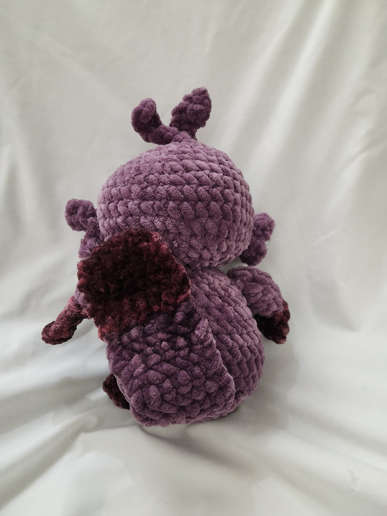 Pictured is a crochet plush of Ralphypusvt , who is a platypus vtuber, from behind. It is made out of two types of purple yarn and custom painted eyes to match their streaming model.