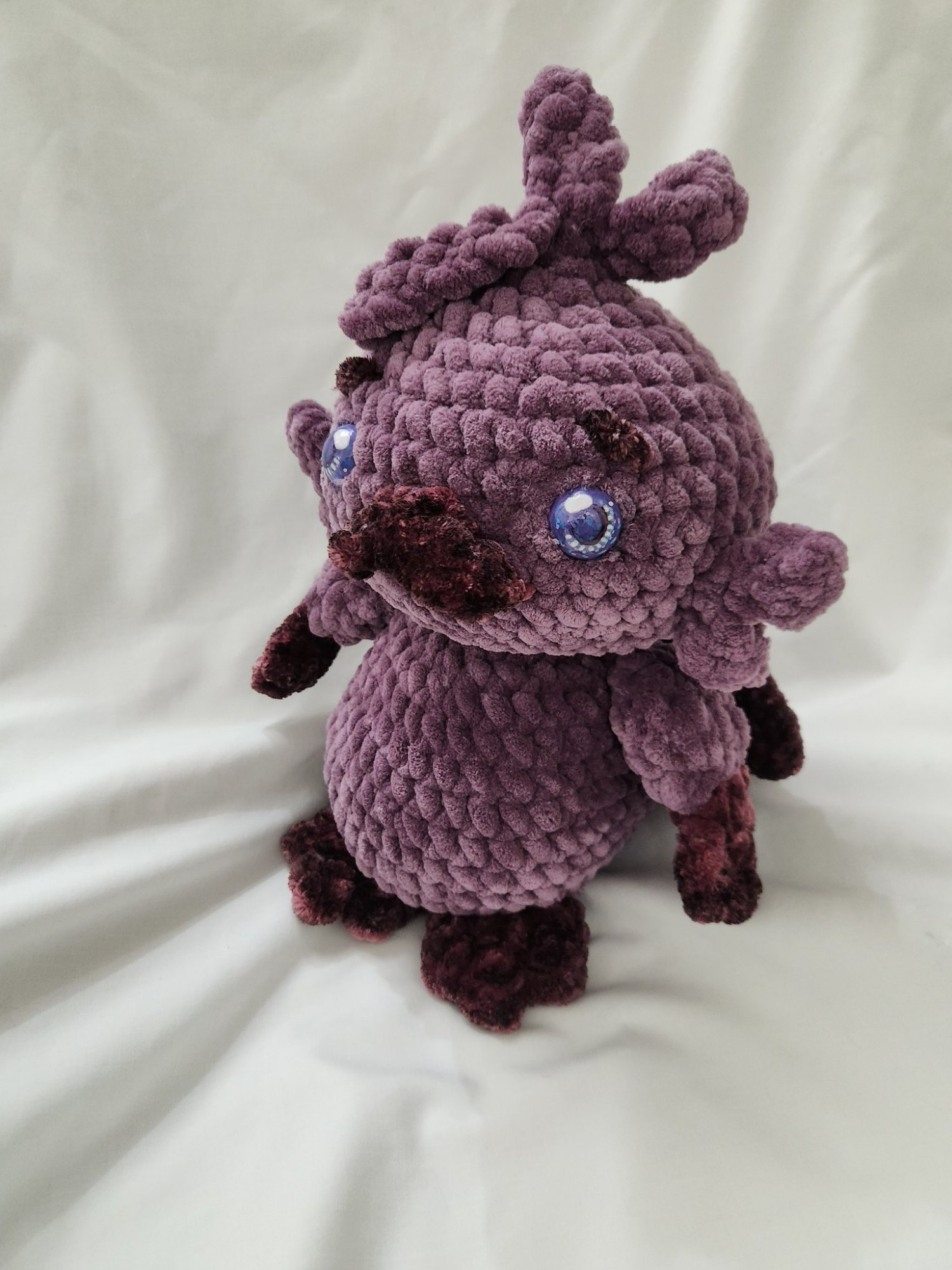 Pictured is a crochet plush of Ralphypusvt, who is a platypus vtuber. It is made out of two types of purple yarn and custom painted eyes to match their streaming model.