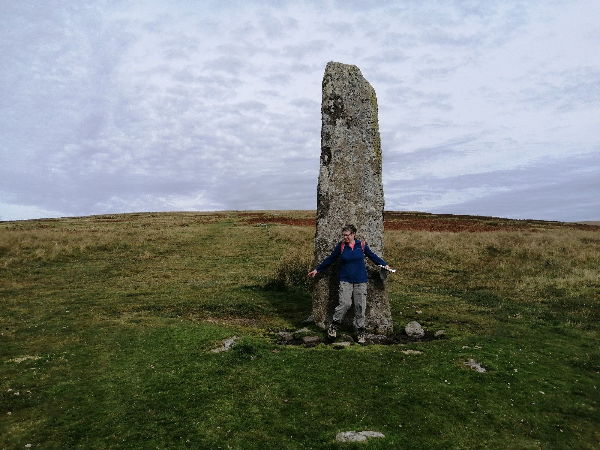 Grey granite standing stone on moorland. Green green grass and grey sky. Person standing in front in blue fleece and grey trousers.