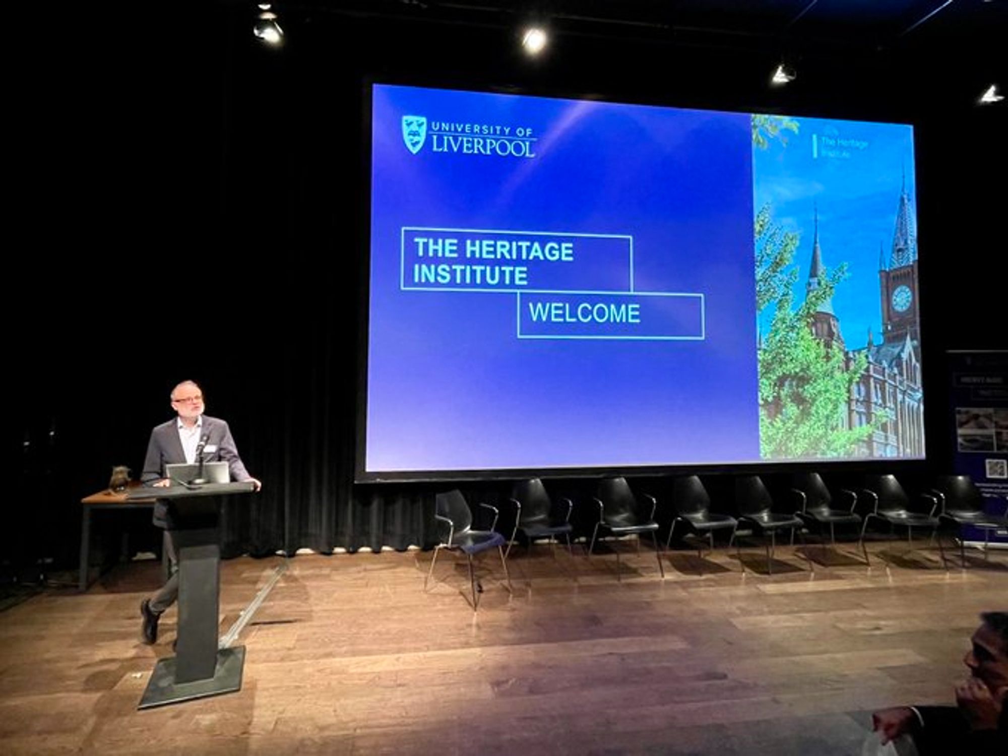 A man making a welcome speech with a screen displaying a slide with 'The Heritage Institute: Welcome' on it