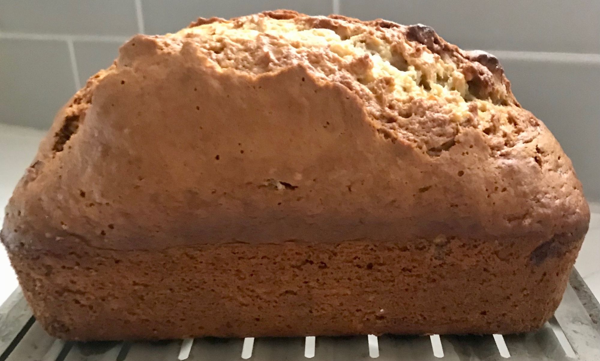 A fresh-baked loaf of banana bread resting on a cooling rack with gray backsplash tiles in the background.