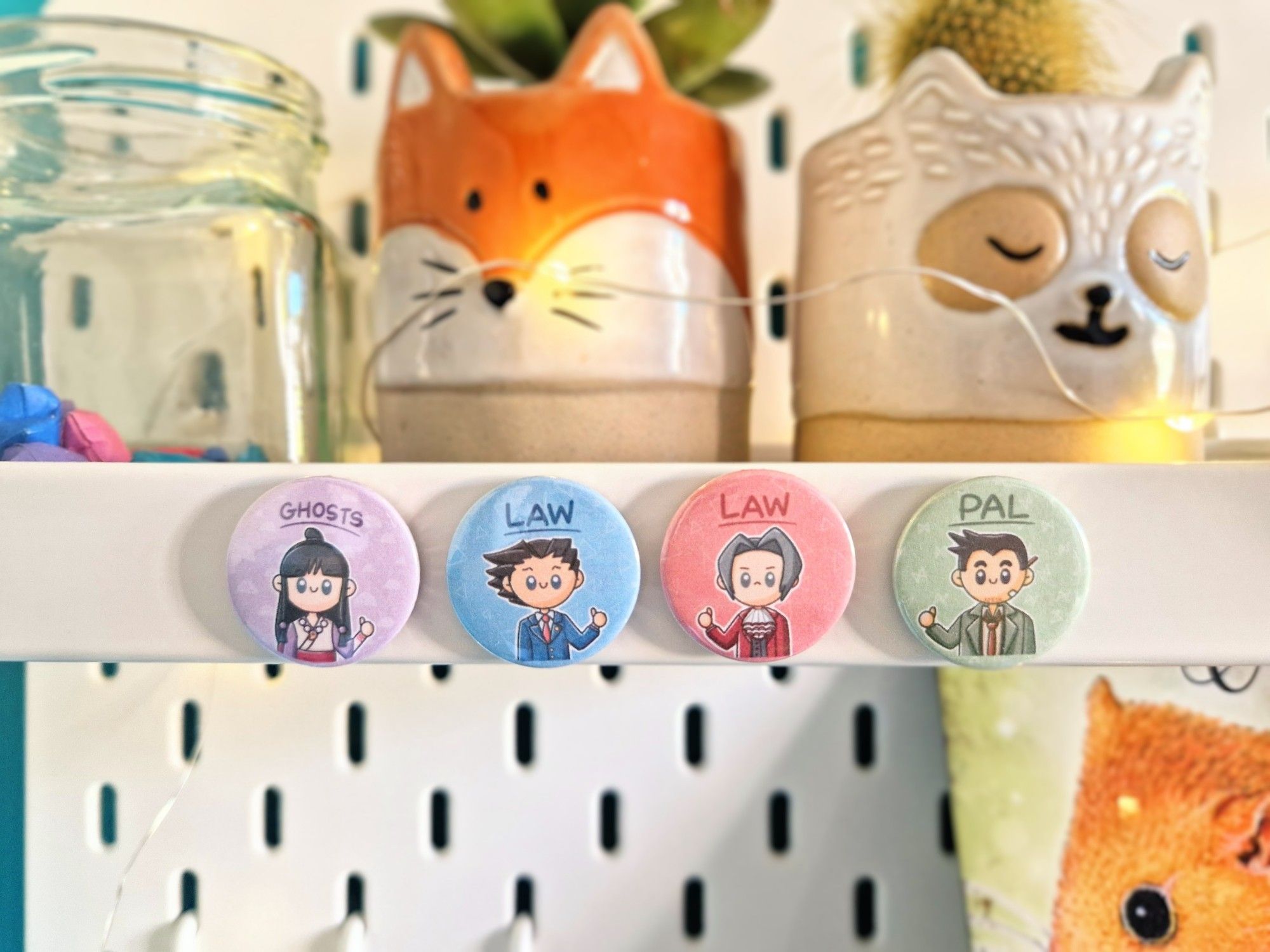 A photo of a small shelf with four round magnets stuck to it. They depict four characters from Ace Attorney in a kawaii style giving a thumbs-up with text above. Maya Fey is saying GHOSTS, Phoenix Wright and Miles Edgeworth are both saying LAW, and Detective Gumshoe is saying PAL. There are some cute animal plant pots and fairy lights in the background.