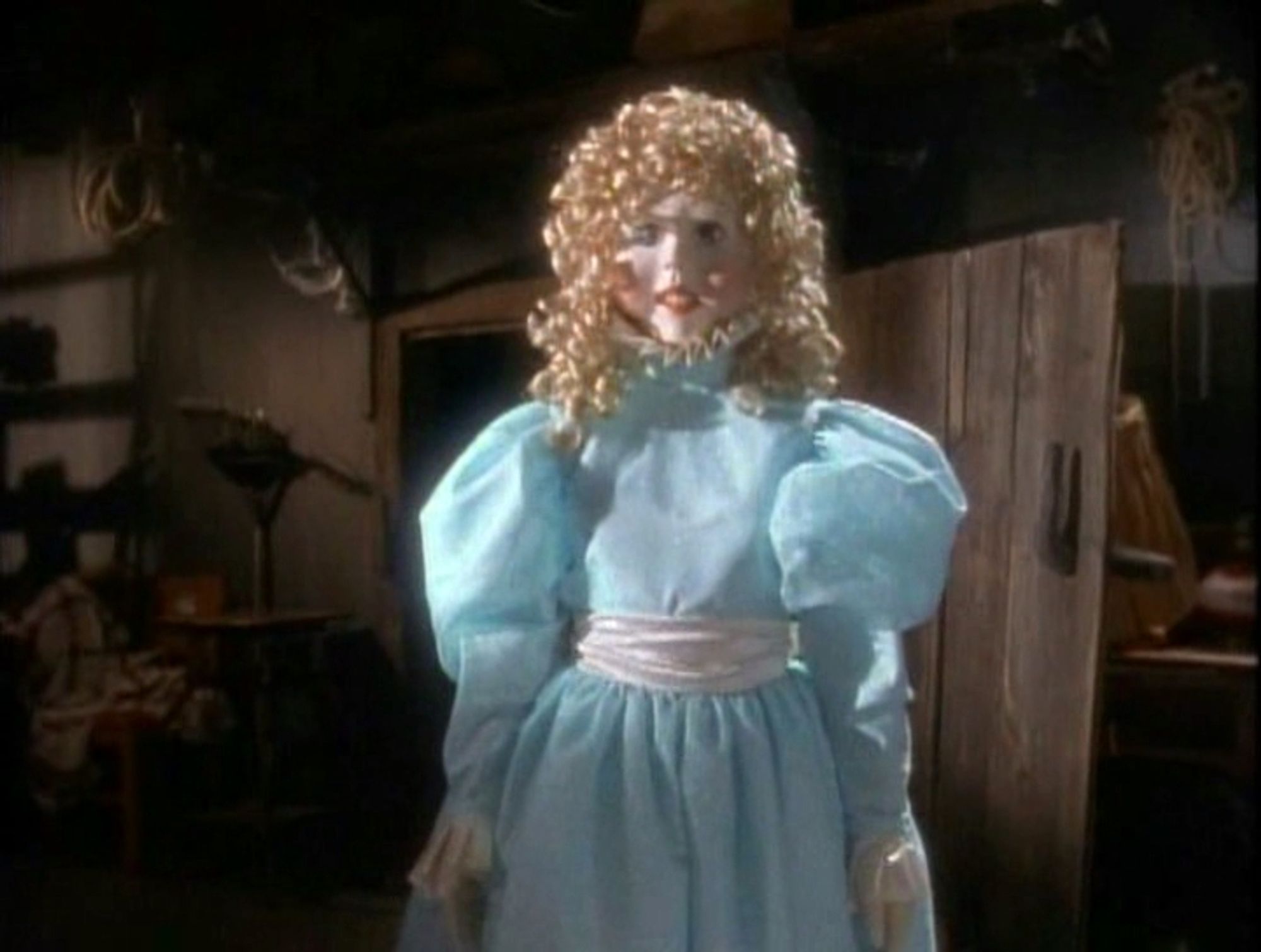 A creepy, life-size doll with yellow curly hair, a light blue princess gown, and white gloves. She is emerging from a creepy wooden door in a basement.