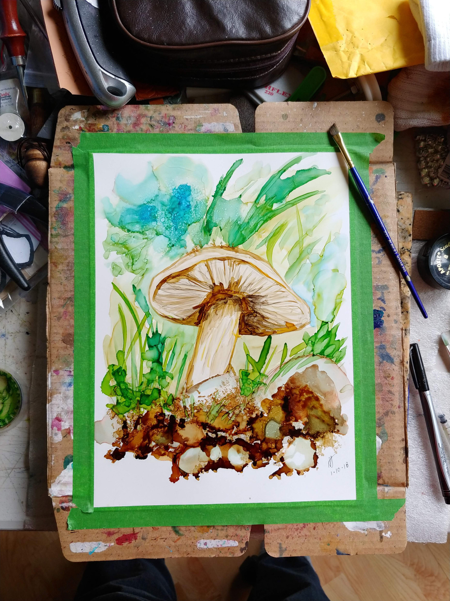 An 8-10 alcohol ink painting taped to cardboard. The painting is a close up of a brown amanita mushroom, dark swells and fluid shapes making up the soil and mulch around it, The angle is from the soil up, to see the gills of the mushroom, and all is flanked by the green of other vegetation.