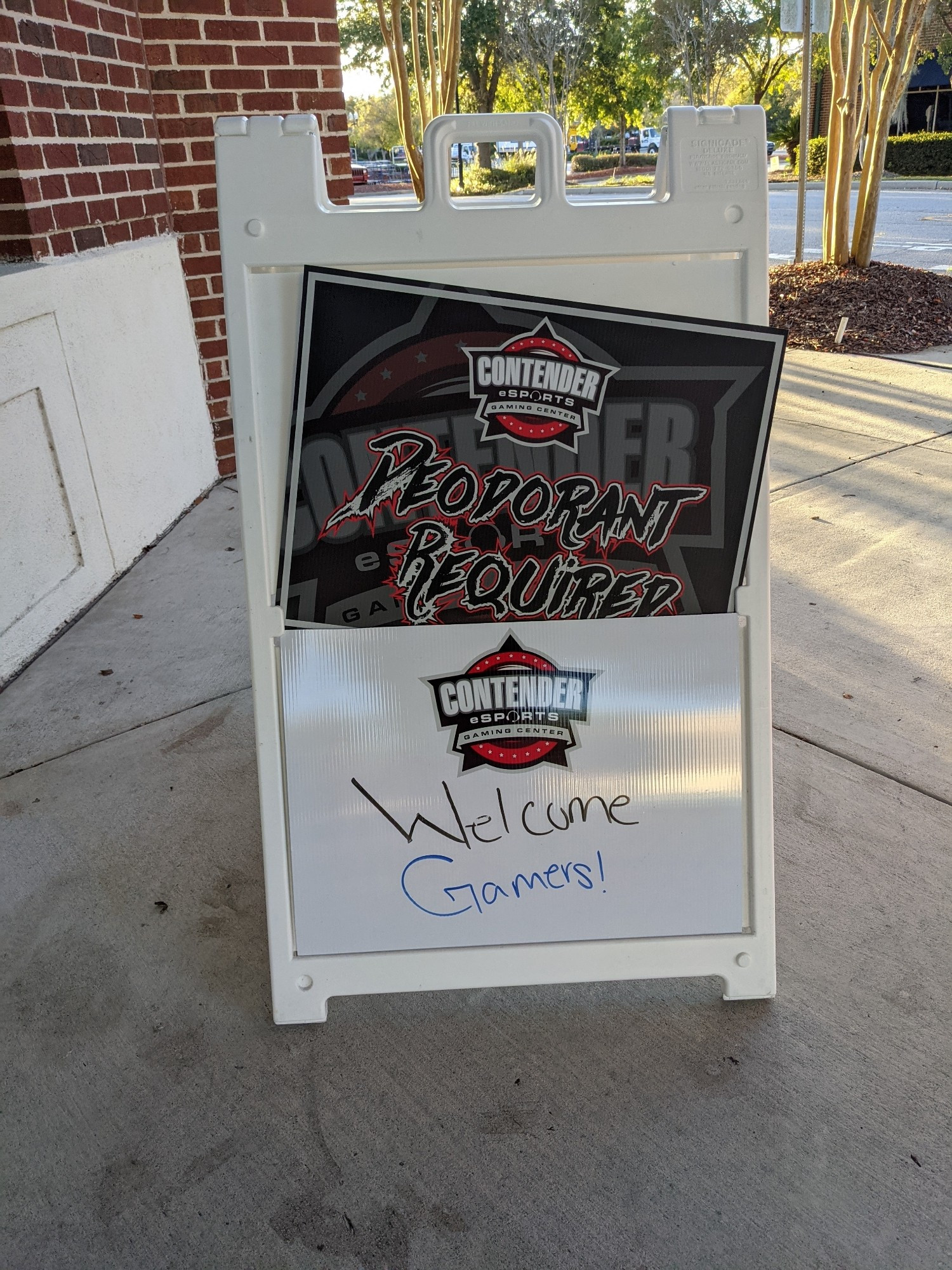 A sandwich board sign with two sections, one with the name of the esports center name and "Deodorant Required" written in "extreme font" and the other a white board with "Welcome Gamers!" handwritten on it.