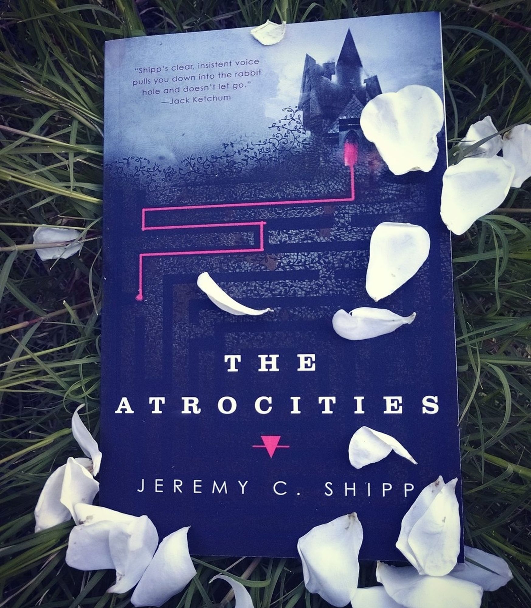 This is a photo of a paperback copy of The Atrocities by me (Jeremy C. Shipp). The book is resting on grass and there are white petals strewn about on the grass and on the cover. The cover features a creepy old manor house, storm clouds, and a hedge maze.