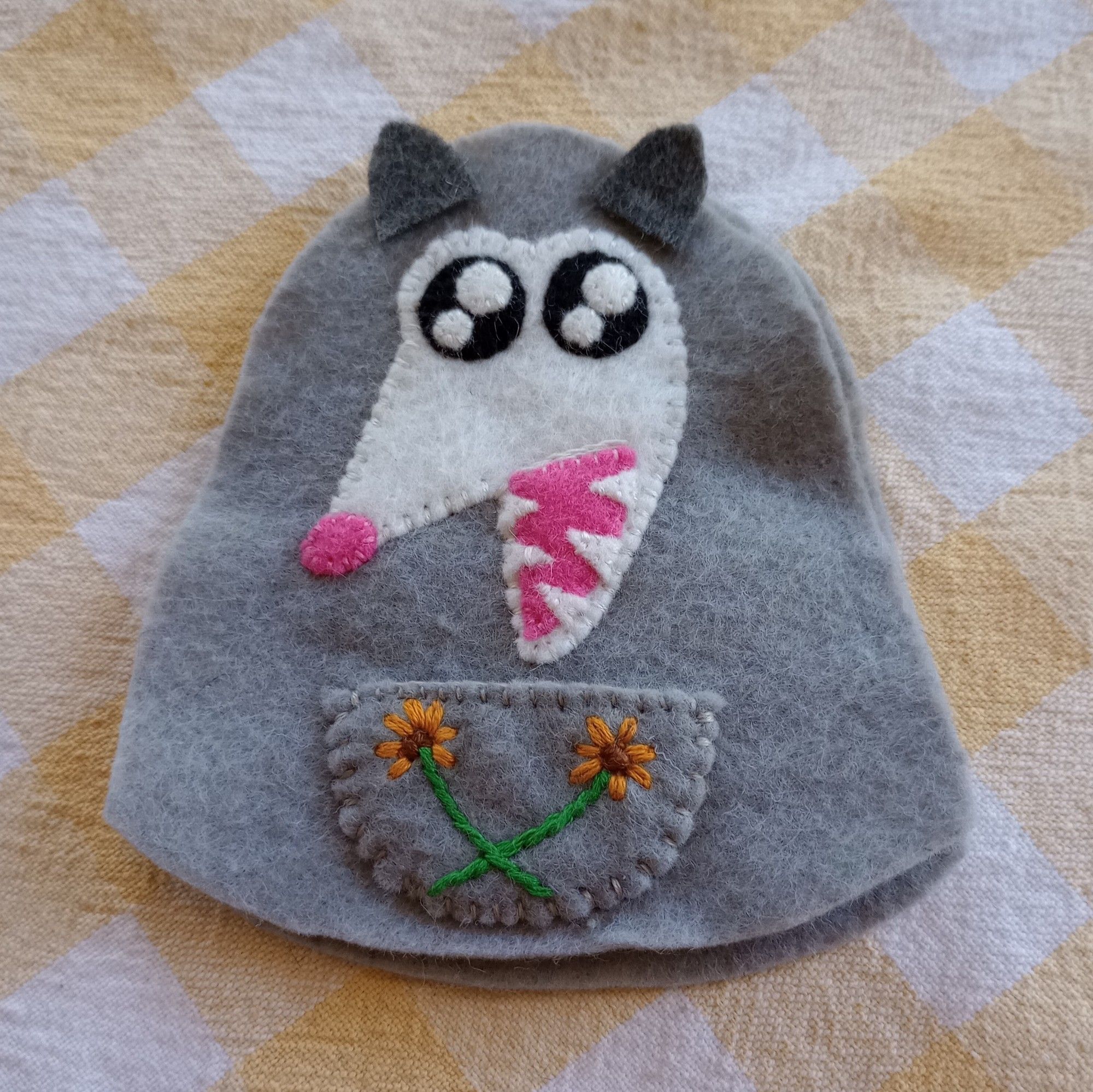 Here's a fabric art bag in progress. She's a gray possum with a white face. She's opening her mouth showing her teeth. She also has a pouch on her belly embroidered with two orange flowers.