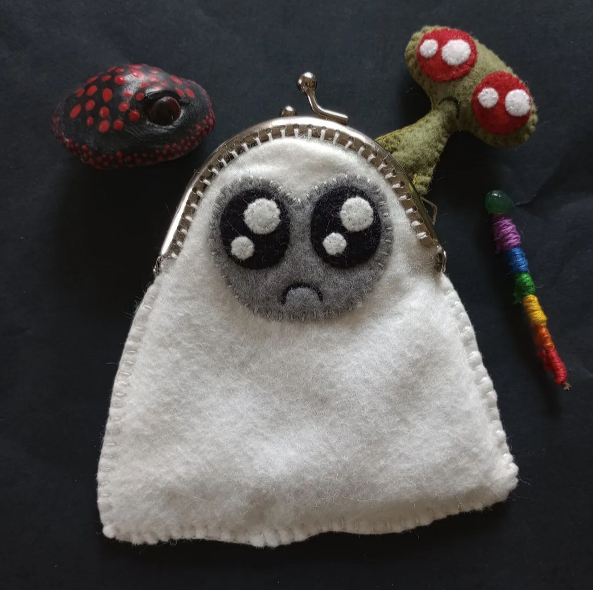 Here's a cute little fabric art bag in the shape of a ghost with a silver coin purse clasp on the top of her head. The ghost is white with a gray face and sparkly black eyes and a little frown. There's a black and white clay mushroom near her with one eye. There's also a little fabric homunculus coming out of her. The homunculus is green with sparkly red eyes and a little frown. There's also a rainbow-colored magical staff beside her with a green stone on top. All of these items are tiny and could be held in the ghost.