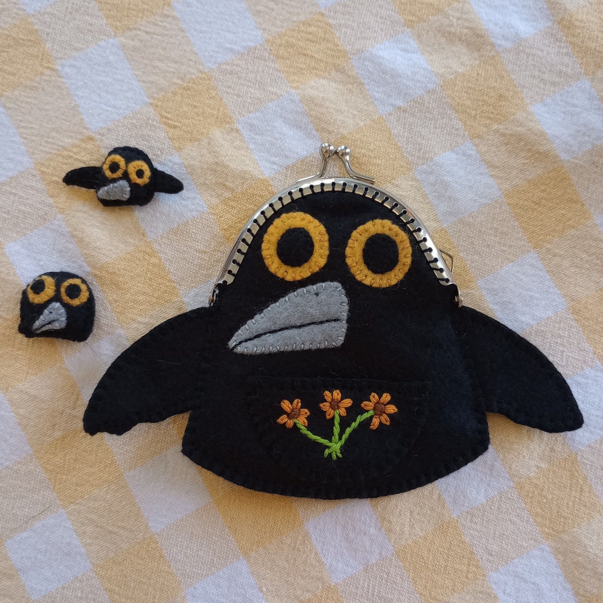 Here's a photo of a fabric art crow bag. She's a cute gumdrop-shaped crow with big yellow eyes and a gray beak. She has a black pouch on her belly with three lovely orange flowers embroidered on it. Beside her are two tiny fabric art baby crows. They also have yellow eyes and gray beaks. One baby has little wings and one has none.