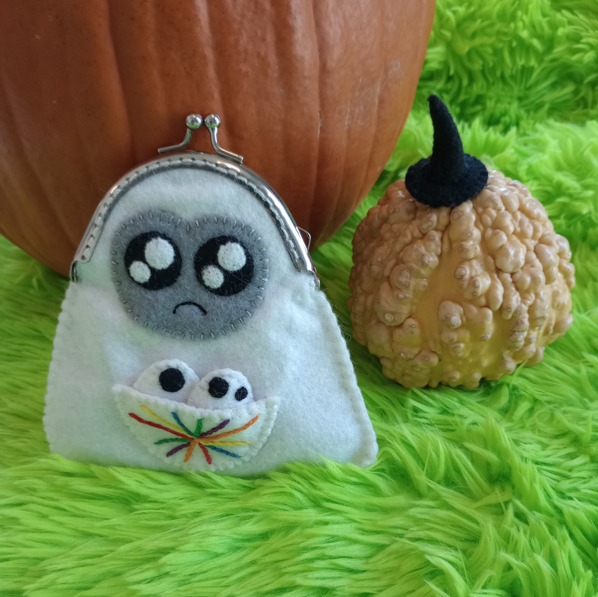 Here's a photo of a fabric art ghost bag with a pumpkin and a decorative gourd in the background. The ghost is white with a gray face and sparkly black eyes. She has a little frown. A pouch over her belly is embroidered with a rainbow star burst. Two little fabric art ghost babies are peeking out of the pouch.