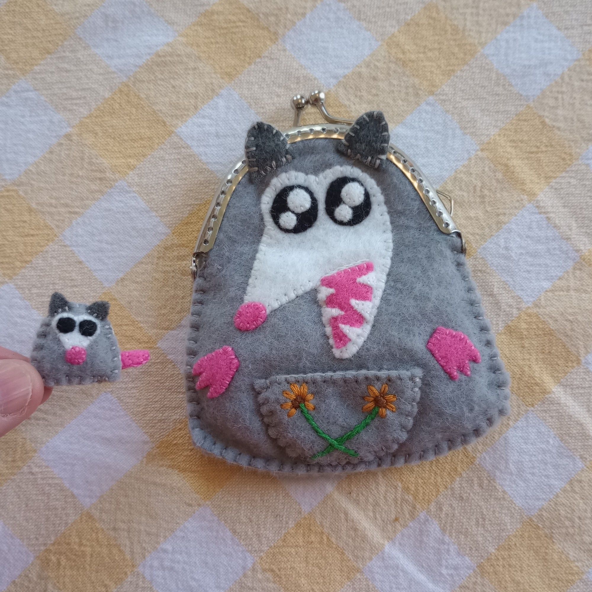 Here's a photo of a fabric art possum bag. She's gray with a white face. She's yelling and showing her sharp teeth. She has a pouch on her belly embroidered with two orange flowers. I'm holding a little fabric possum baby between two fingers. The baby has two black eyes and a little tail.