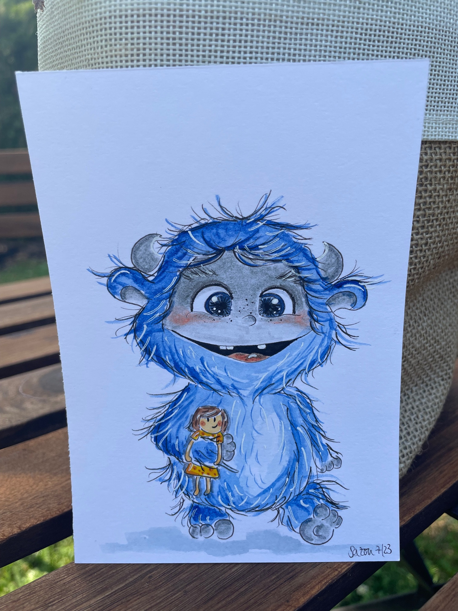 Blaues wuscheliges Monsterchen mit lächelndem Gesicht und offenen Mund, die Zähne und Zunge blitzen hervor. Es hat eine Puppe mit gelben Kleid unter dem Arm geklemmt. Das Bild steht auf einem Holztisch im Garten.