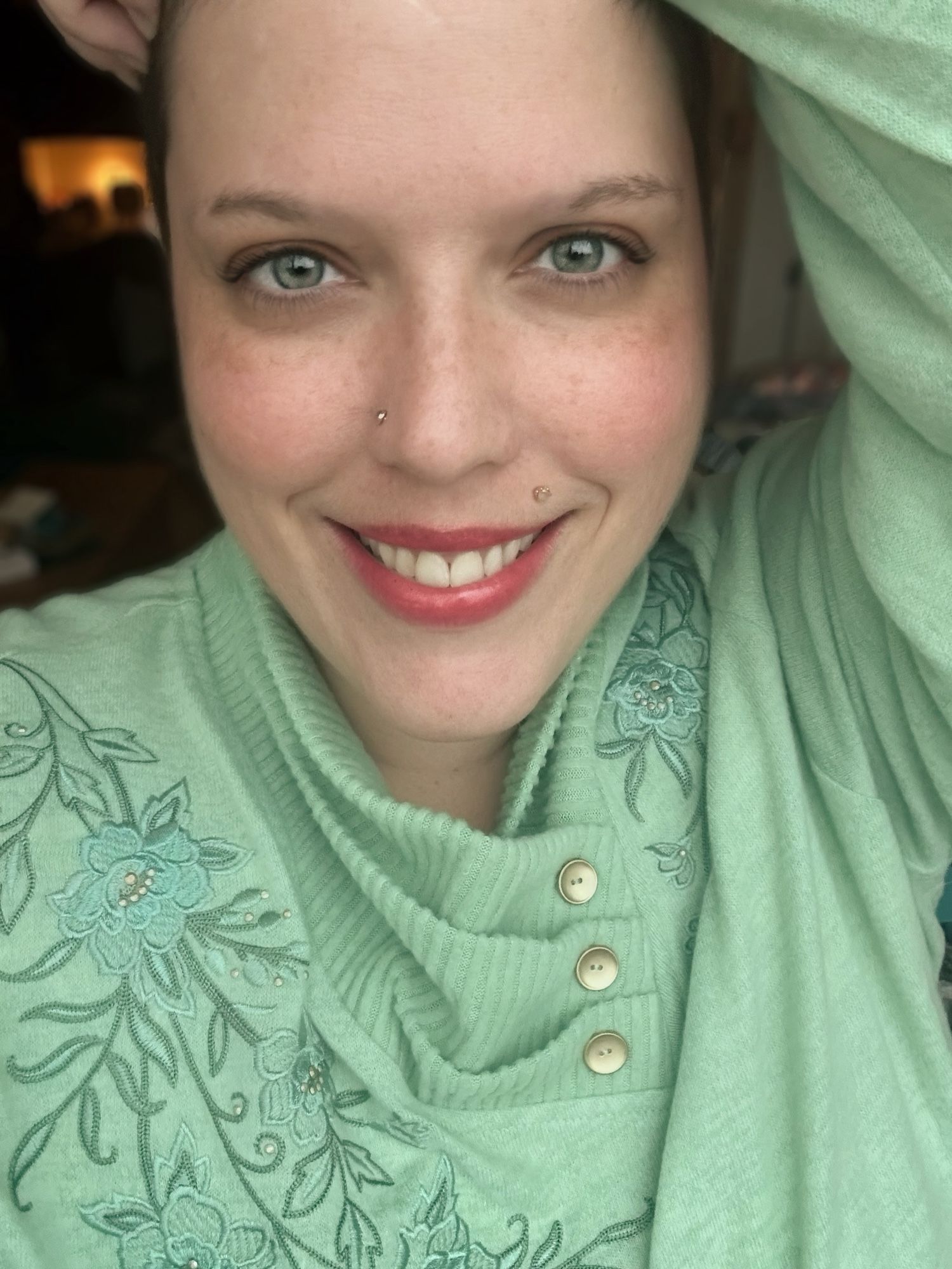 Portrait of a woman smiling. She’s wearing a mint green Christmas sweater with buttons on the neck. Her lips are stained mulberry red and her eyes are green.