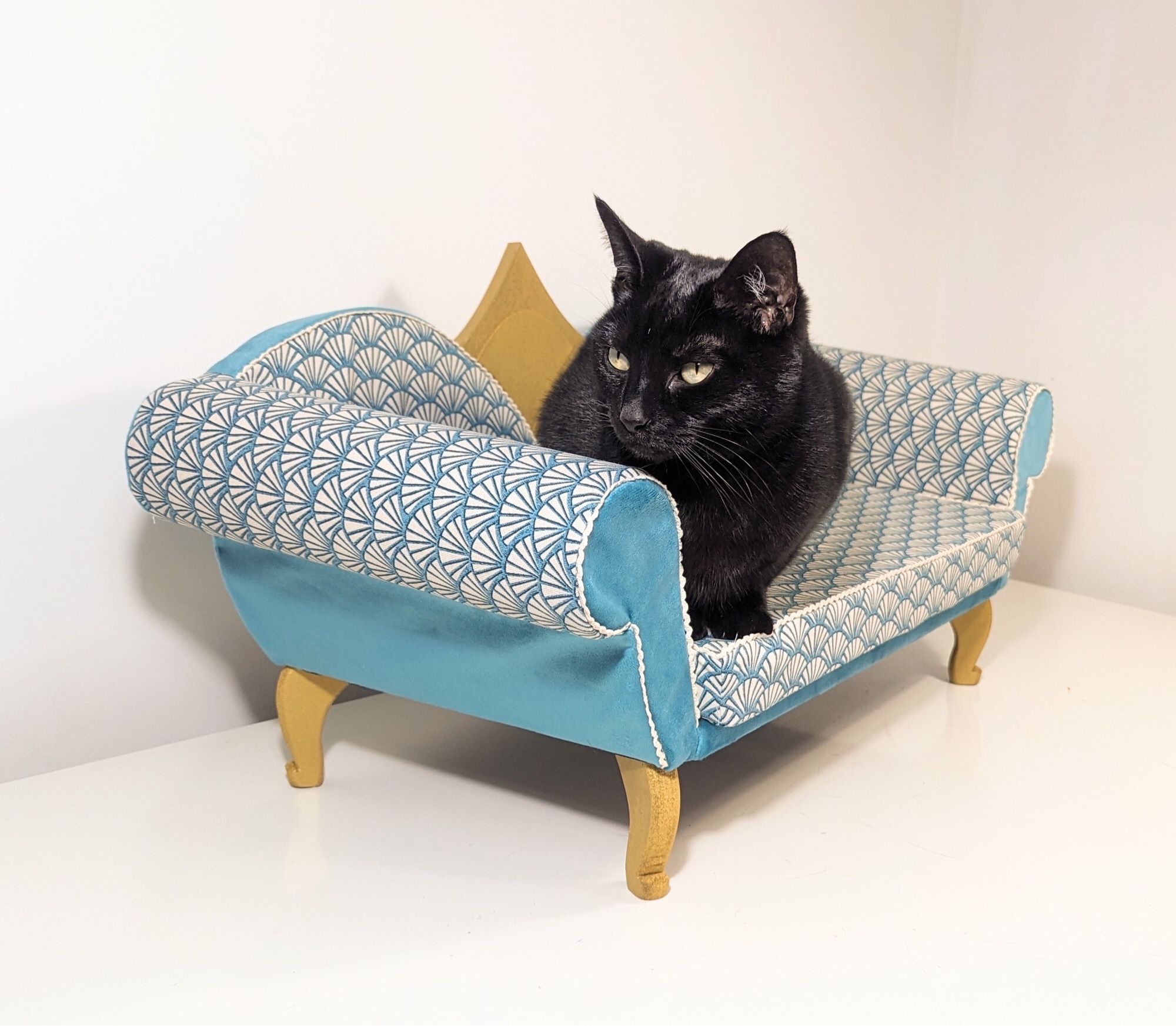 Black cat crouches on a fancy teal/blue Art Deco inspired cat sofa with gold feet and detailing