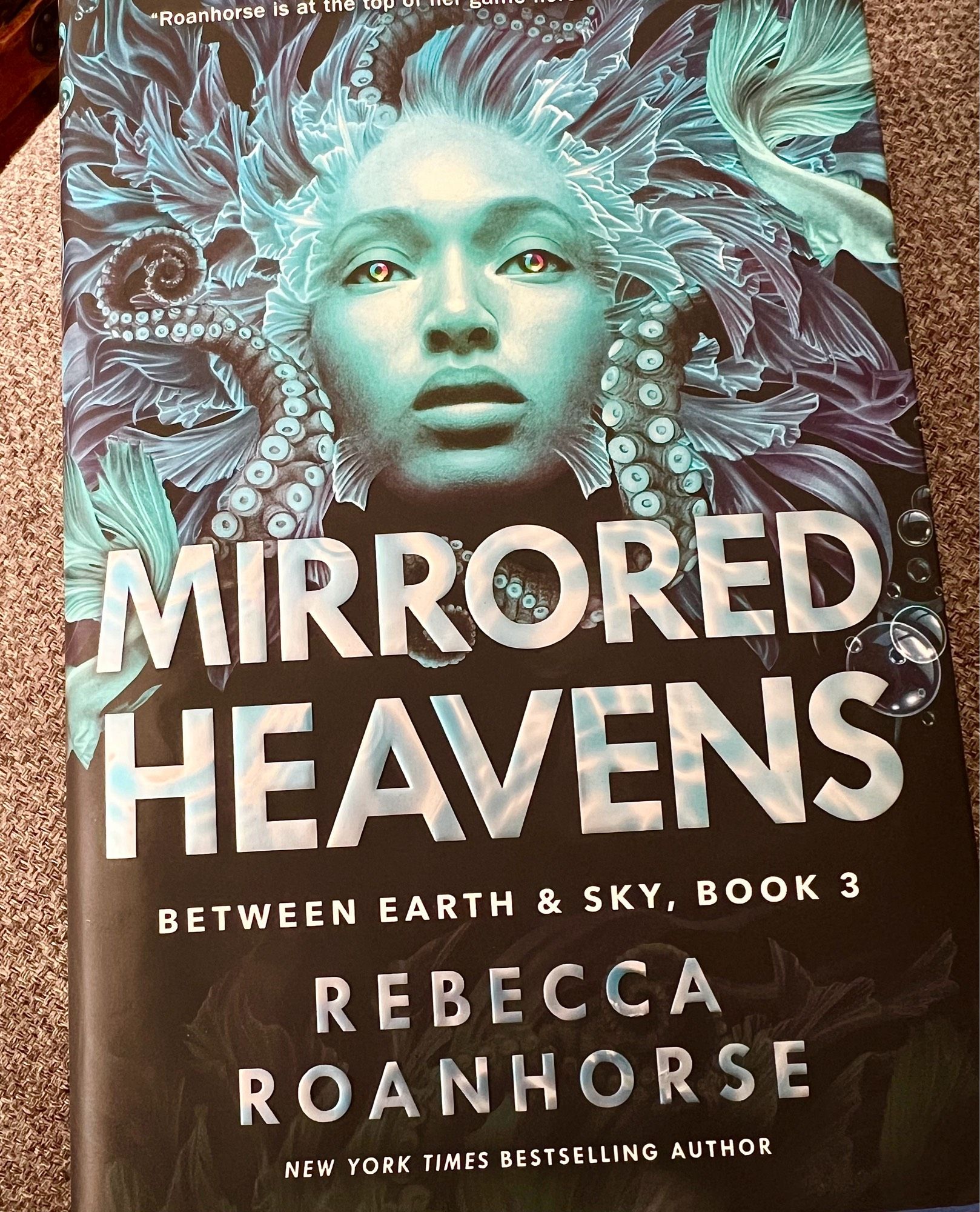 Hardcover “Mirrored Heavens, Between Earth and Sky Book 3” by Rebecca Roanhorse. The cover art is of Xiala, one of the main characters, with fins, tentacles, and other water-y imagery.