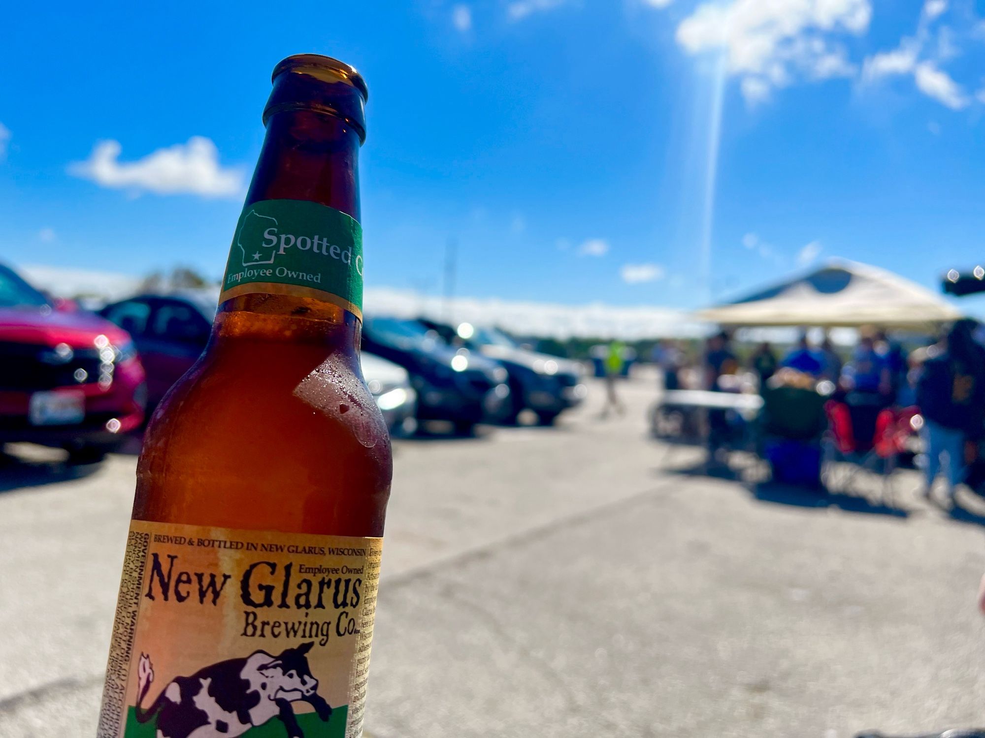 A bottle of Spotted Cow with the parking lot of American Family Field and some tailgating tents in the background.