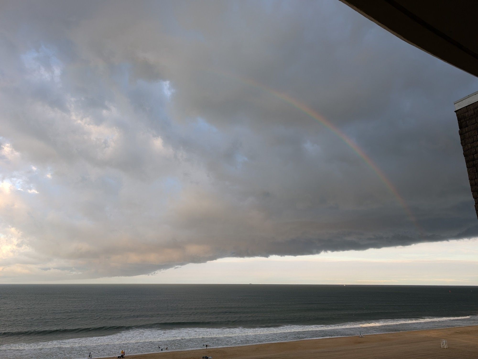 A photo of a somewhat calm beach and a strong front moving through the sky, pushing clouds along in a line and casting a rainbow over the ocean.