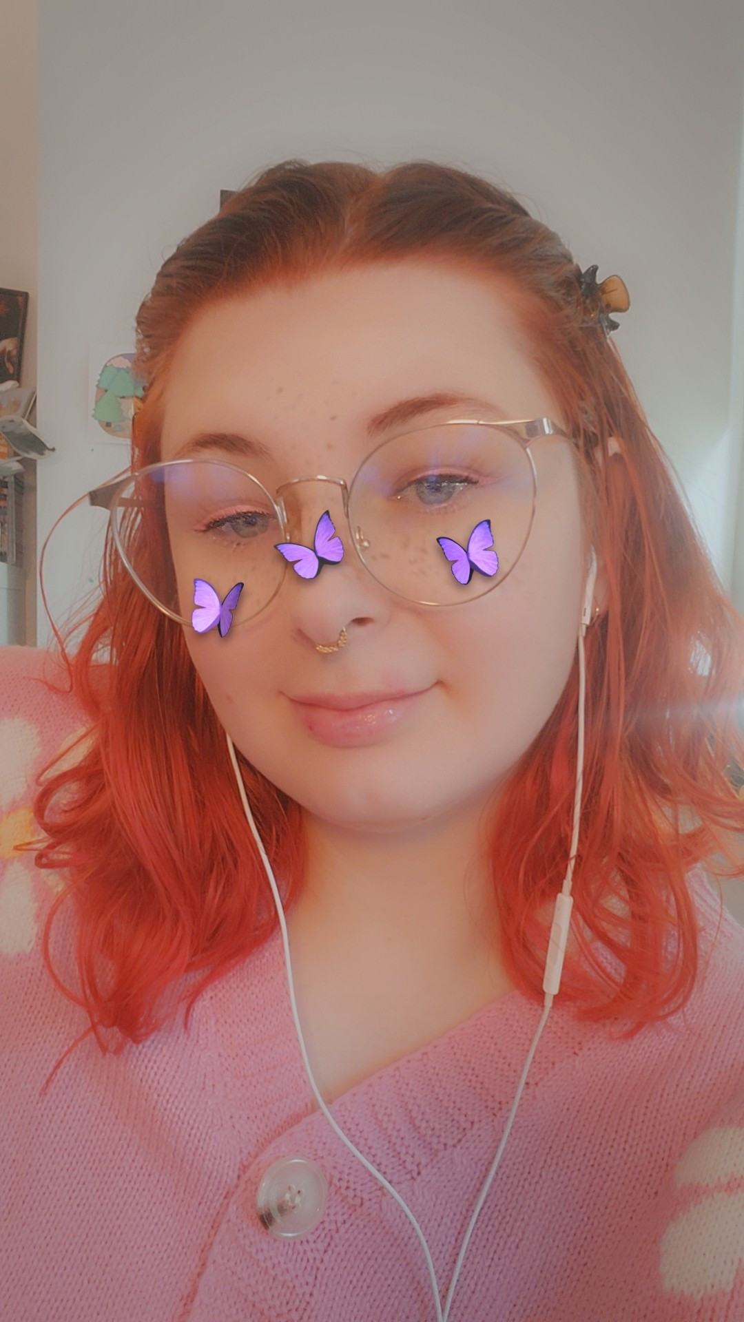 selfie of a white girl with orange hair and glasses, wearing a pink cardigan with a Snapchat filter with three purple butterflies on her face