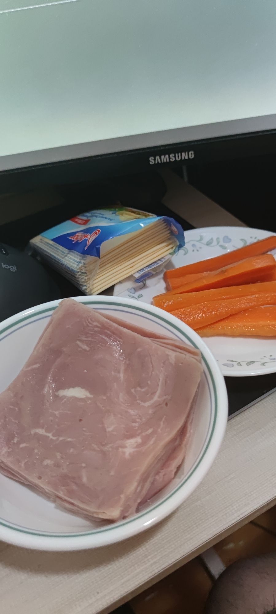 A bowl of sliced supermarket ham, packet of cheese slices, and a half-dozen pickled carrot sticks sitting on my desk in front of the computer