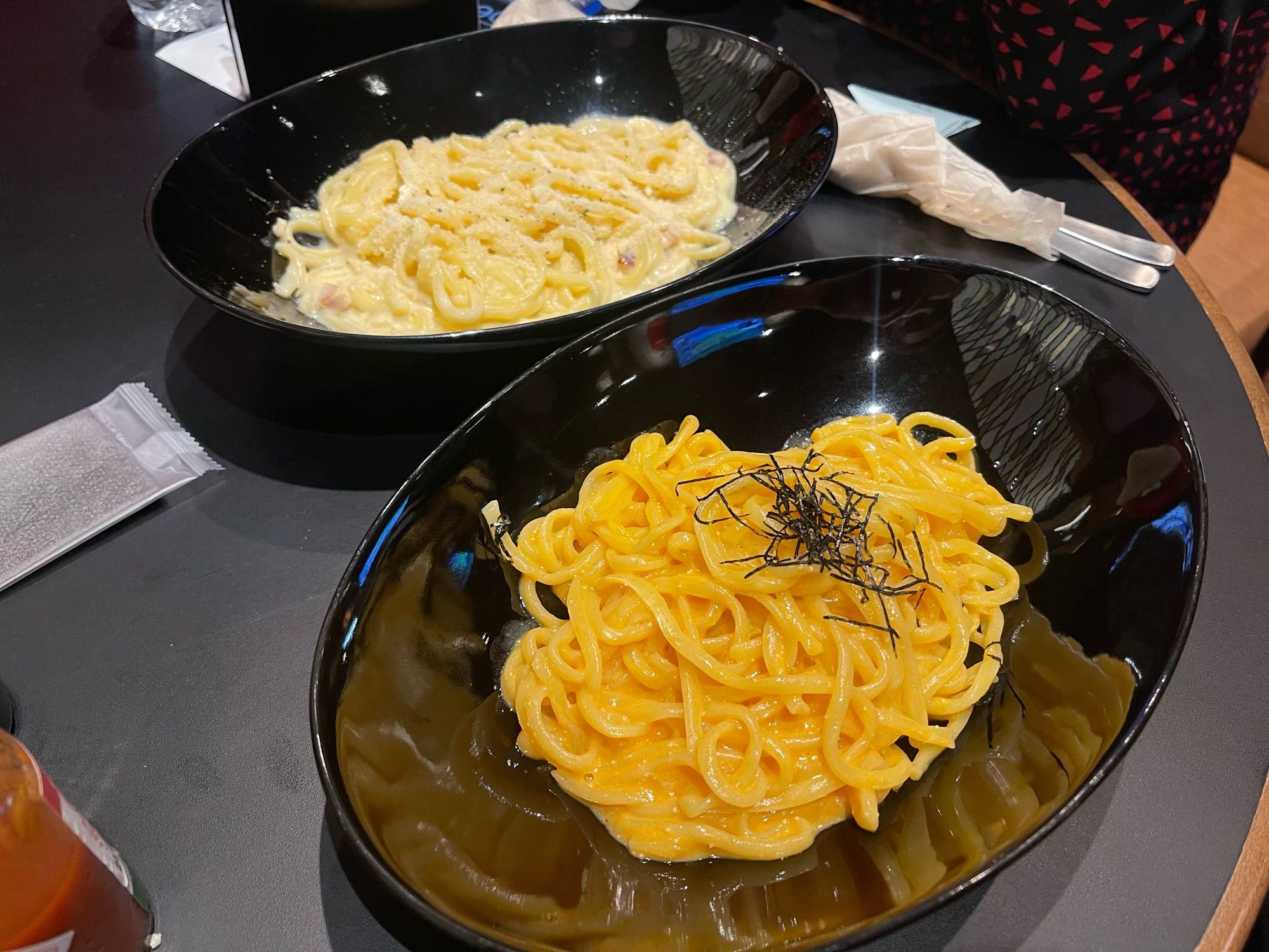 Photo of black shiny plates, one of white creamy carbonara and one of bright orange creamy mentaiko pasta with seaweed strips on top