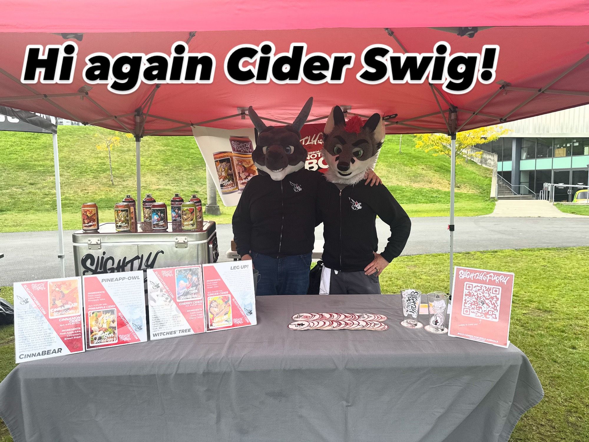 Picture of a vendor booth for Slightly Furry at a cider tasting event with two people wearing a dragon and fox costume head. On the table in front of them are advertisements for different ciders and the brand.