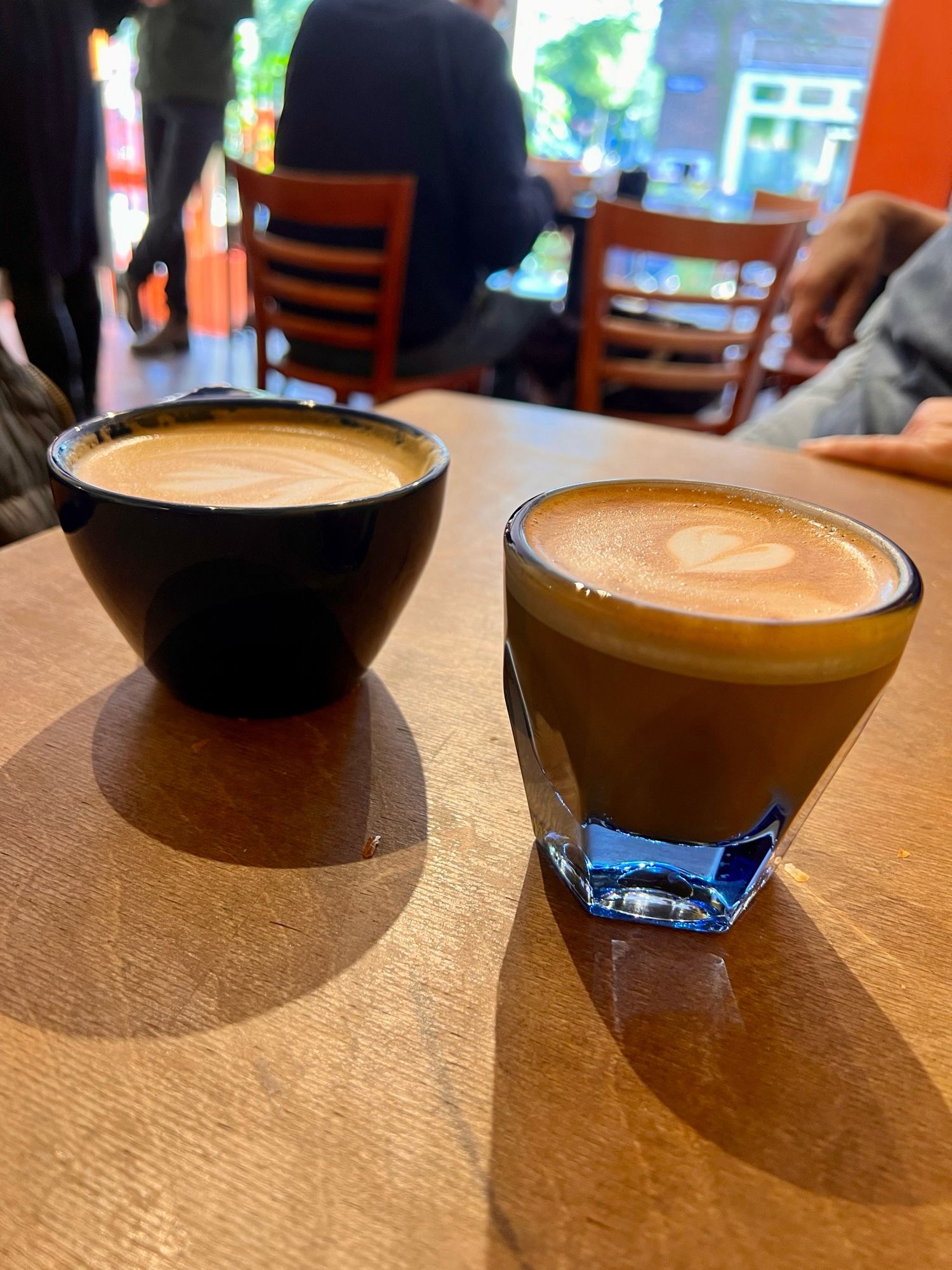 Een glazen kop koffie en een stenen kop koffie. Met schuim en hartjes. Op een bruin tafeltje