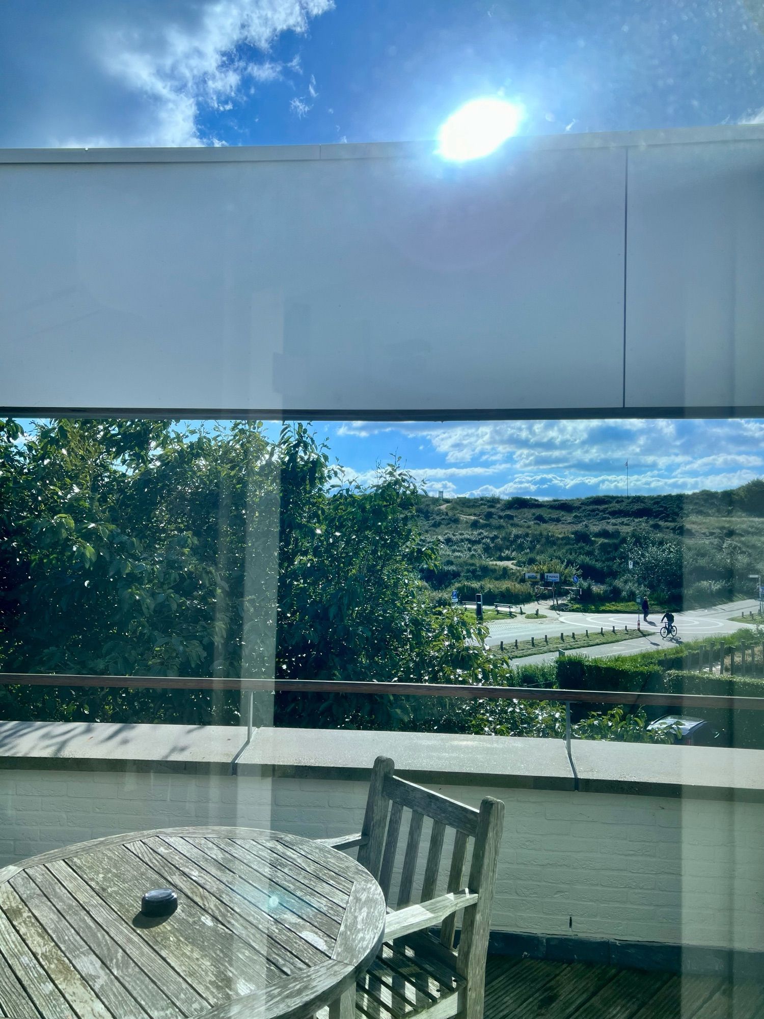 Terrastafel met stoel op een balkon. Erachter uitzicht op de duinen. Blauwe lucht met wolken en een helder schijnende zon