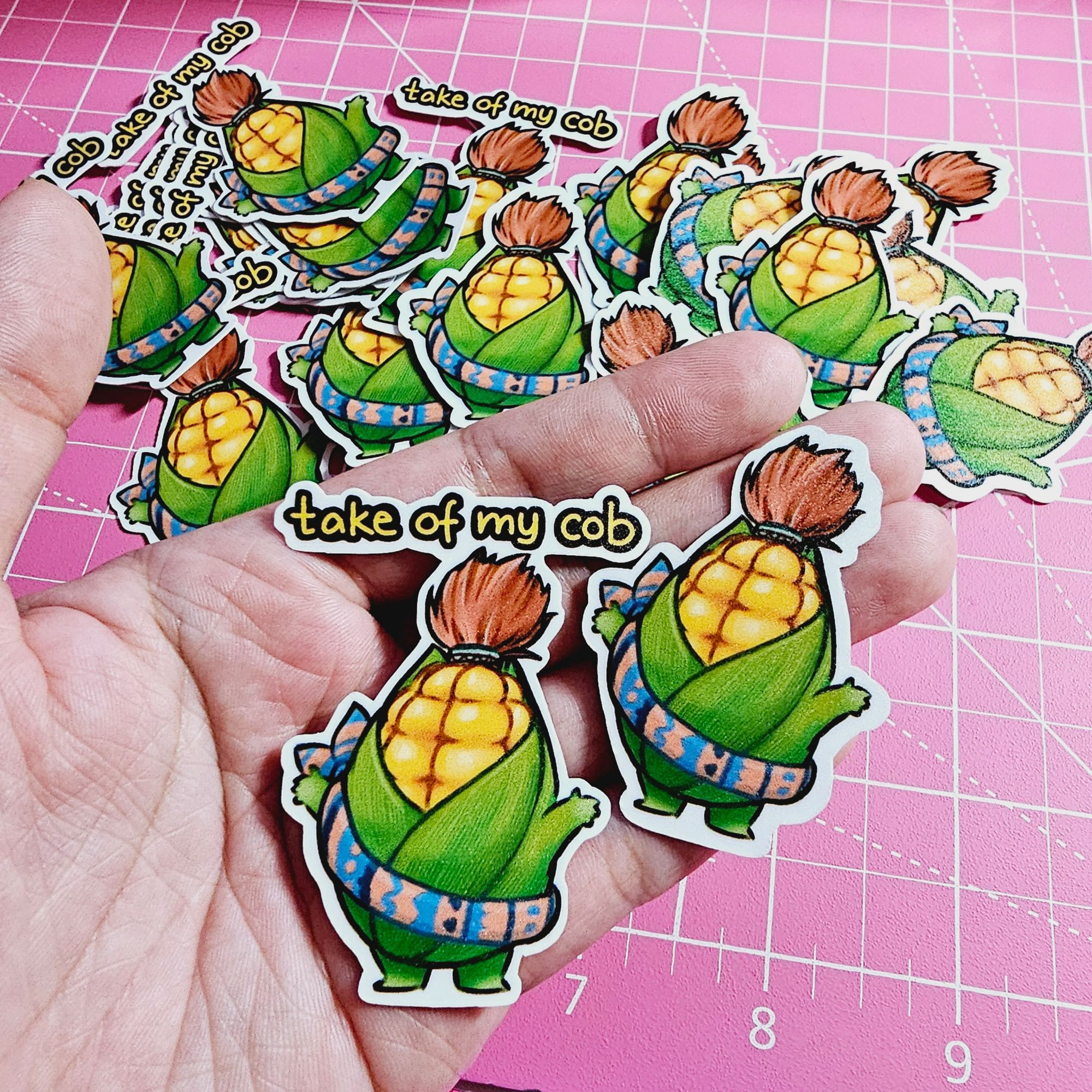 Cornservant stickers I drew for my shop. There are two variants shown in the palm of my hand. On the left is a version of the cornservant with text above it saying "take of my cob" in black-bordered yellow text. The one on the right is the same sticker without the text. In the background are multiples of the two sticker designs scattered on a pink grid craft cutting board.