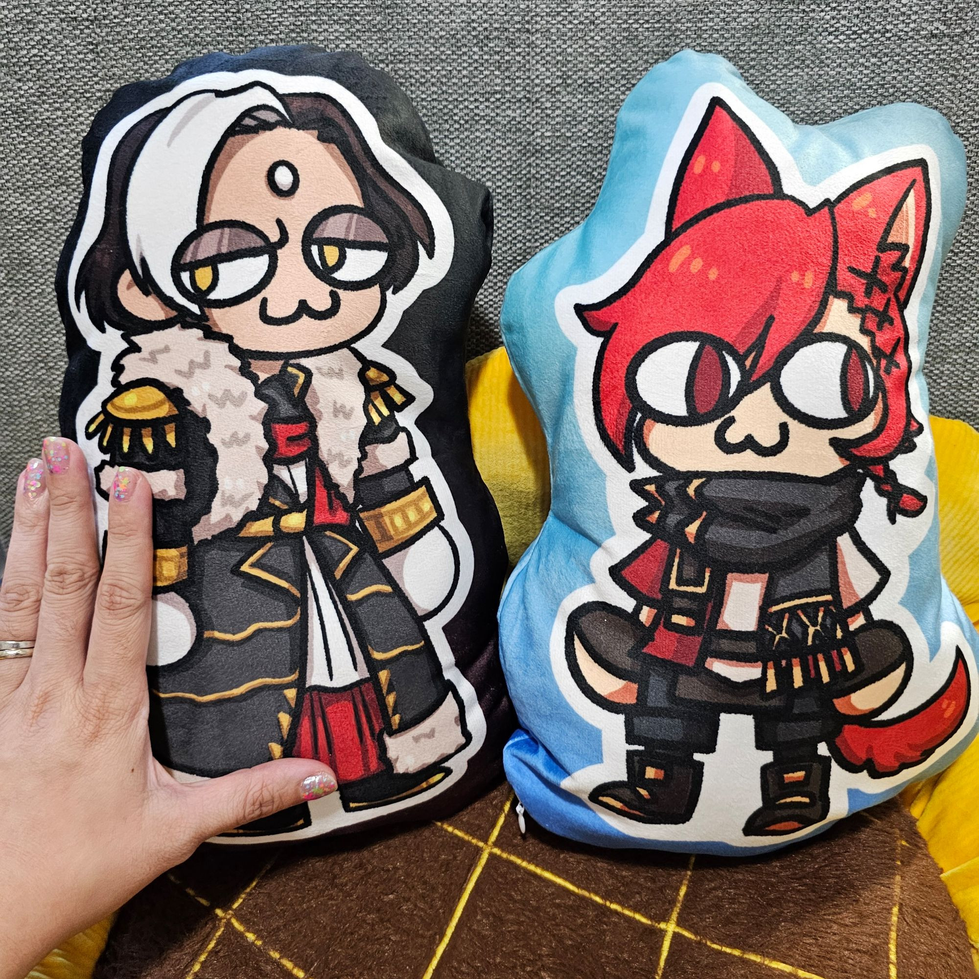 Small pillows featuring two beloved characters from Final Fantasy XIV. They are designed by me and were drawn in the Neco Arc style. There's Emet-Selch and G'raha Tia as shown in Shadowbringers and onwards. The pillows sit on a Sunflower cushion. My hand is also shown for size comparison.