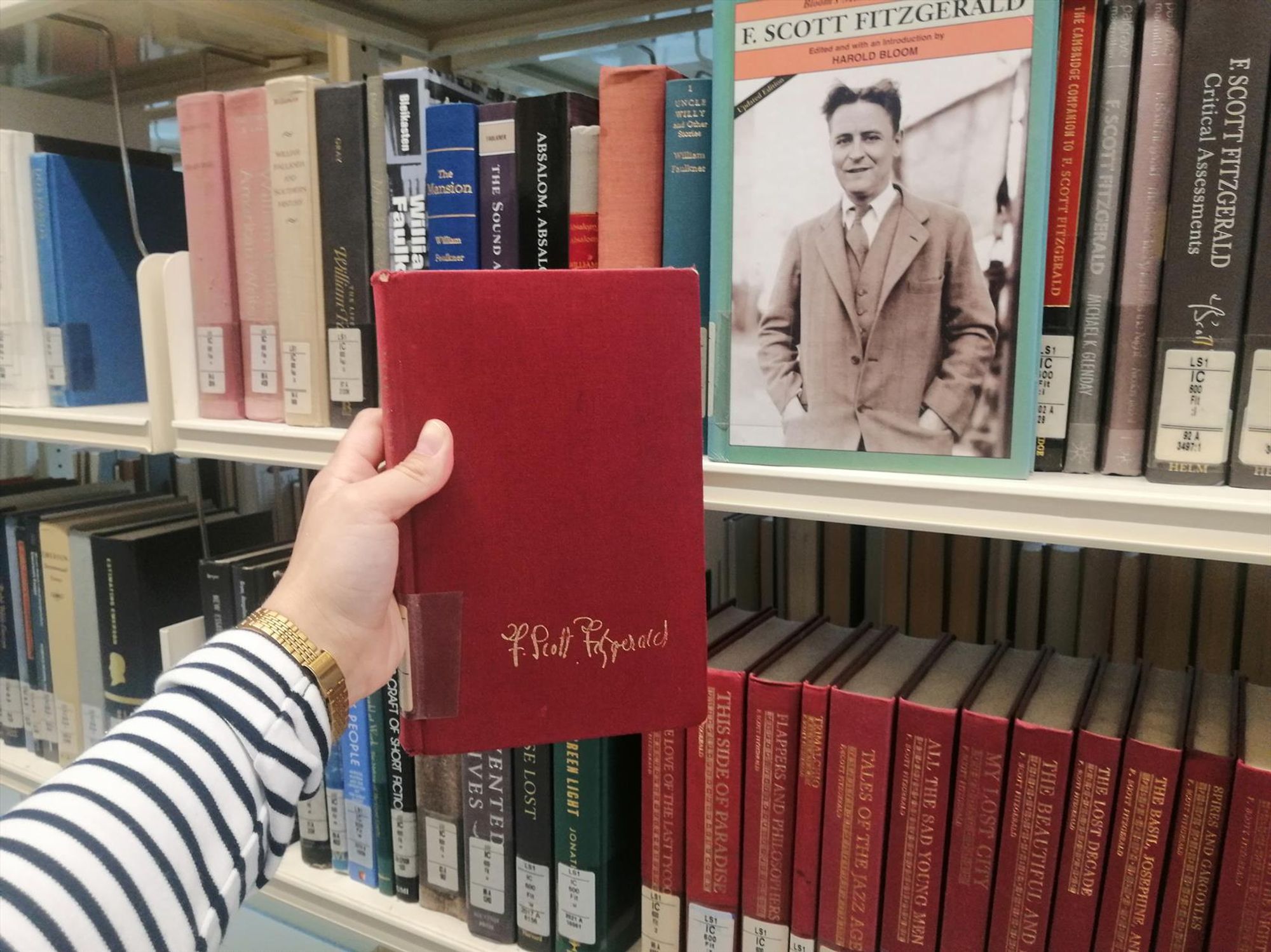 Holding the novel "The Great Gatsby", bound in red cloth with Fitzgerald's signature in gold, in front of bookshelves filled with his works. On the top shelf sits a book with the cover to the front showing a photograph of Fitzgerald