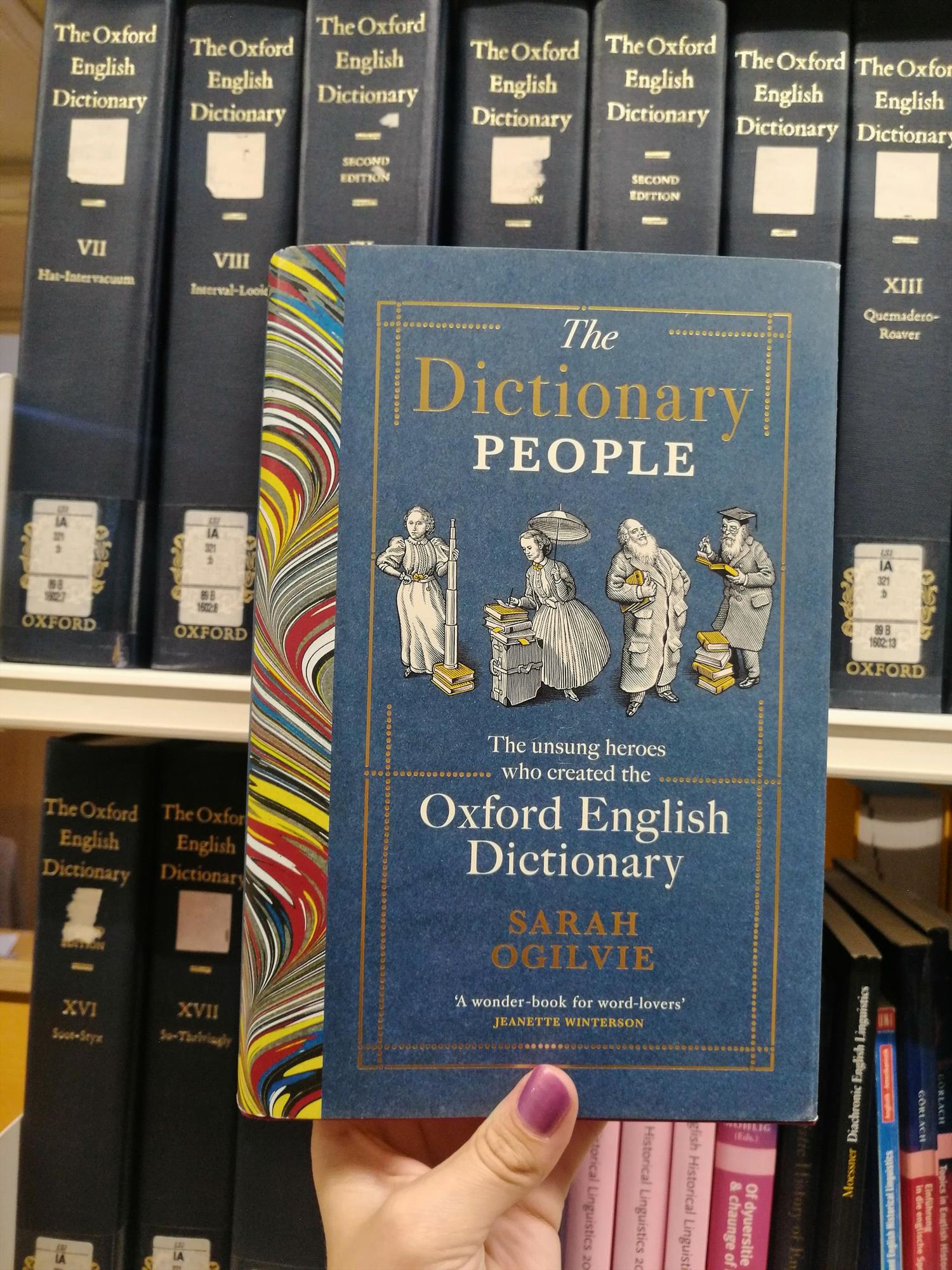 Holding Sarah Ogilvie's book "The Dictionary People" in front of some bookshelves containing the Oxford English Dictionary