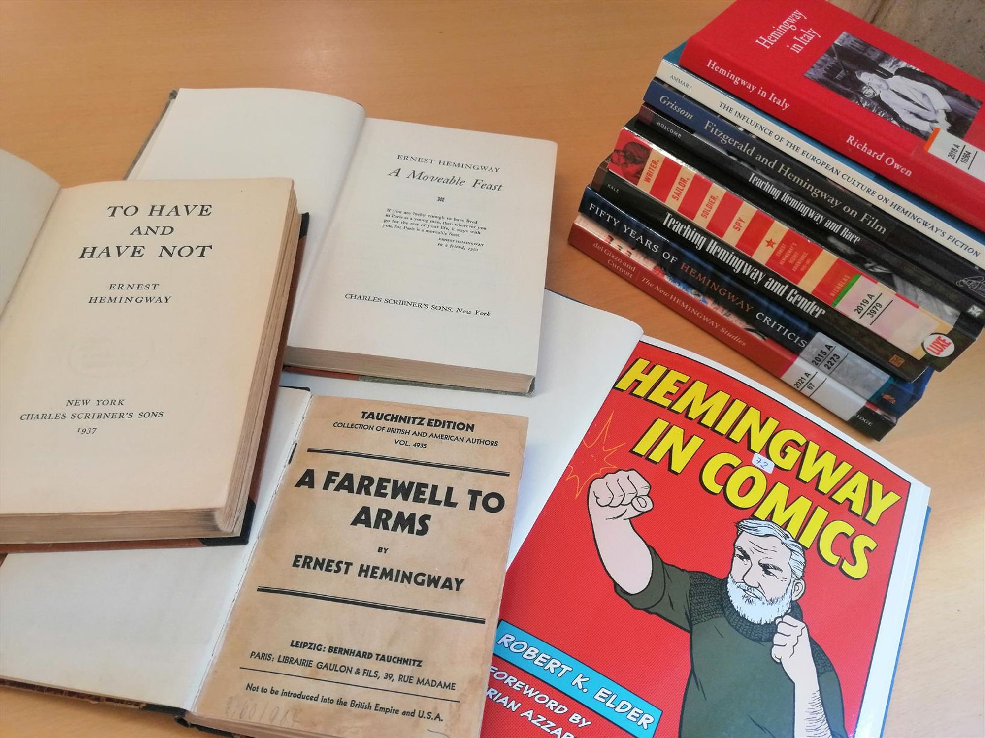  A stack of books of secondary literature on Hemingway, as well as 3 open primary texts by Hemingway, and a large volume called "Hemingway in Comics"