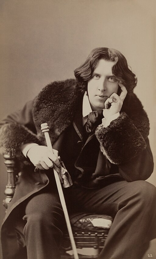 Oscar Wilde, a white man photographed in sepia colours by Napoléon Sarony is sitting on a chair, looking at the camera thoughtfully. He is wearing a suit and a coat with fur collar and holds a cane in one hand