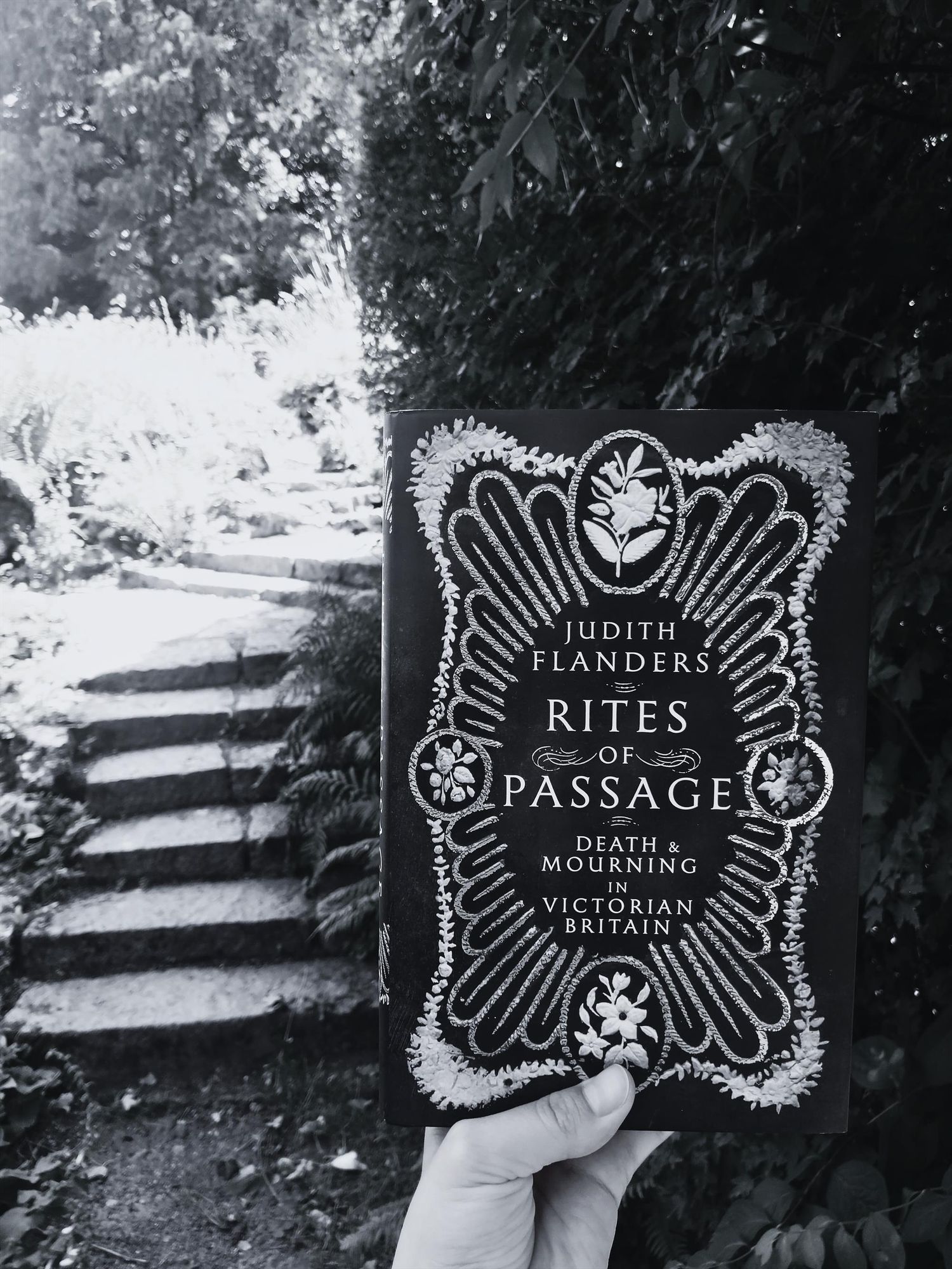 A black and white rendition of a photo holding Judith Flanders's book "Rites of Passage: Death & Mourning in Victorian Britain" in the Botanical Gardens, the background shows stone stairs next to a dark and looming wall of ivy