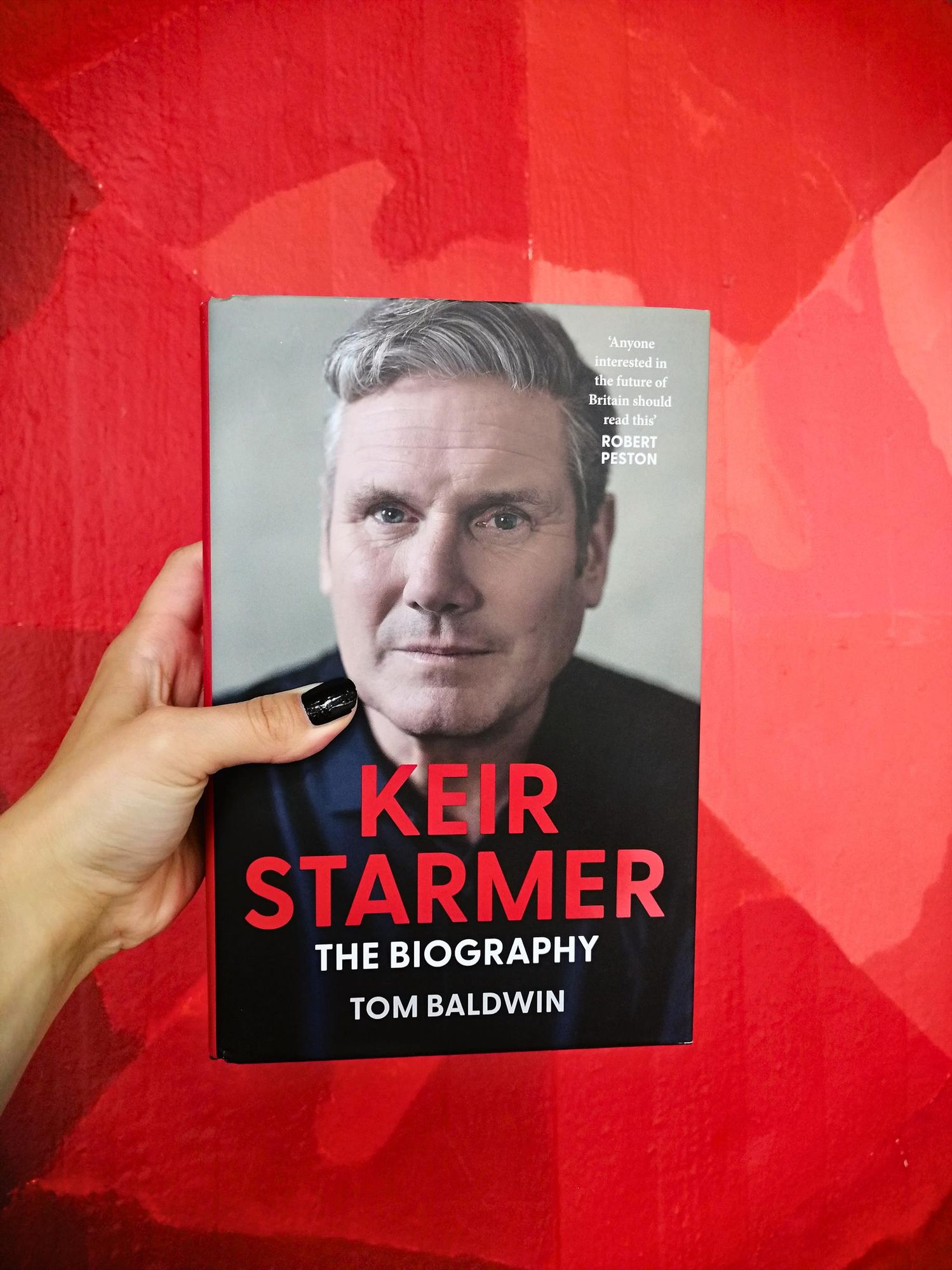 Holding the biography "Keir Starmer" by Tom Baldwin in front of a red poster