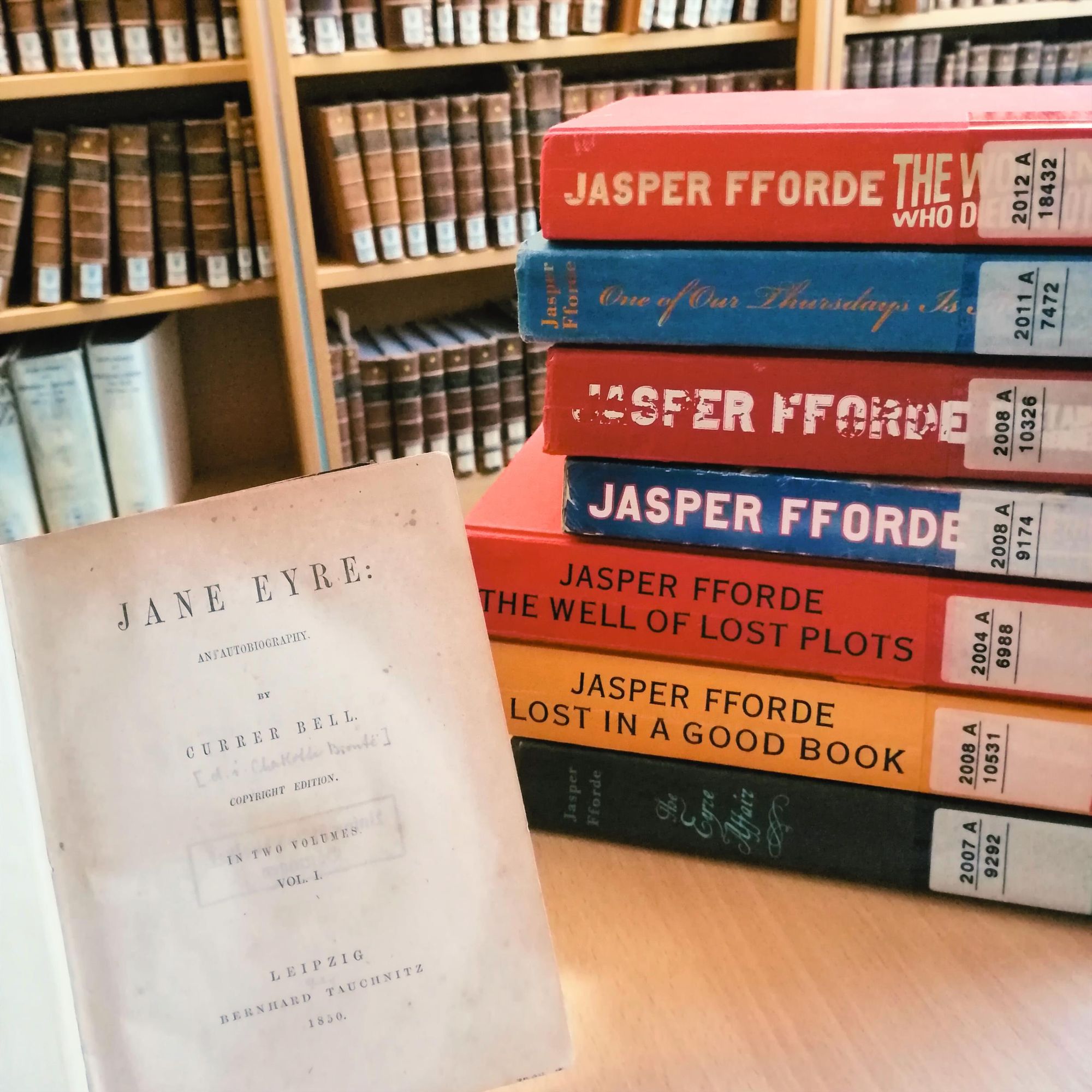 in front of some tall bookshelves filled with old, brown, leather-bound tomes is a wooden table, on which are placed: on the right a stack of the books of Jasper Fforde's "Thursday Next" series, and on the left an 1850 Tauchnitz edition of "Jane Eyre"