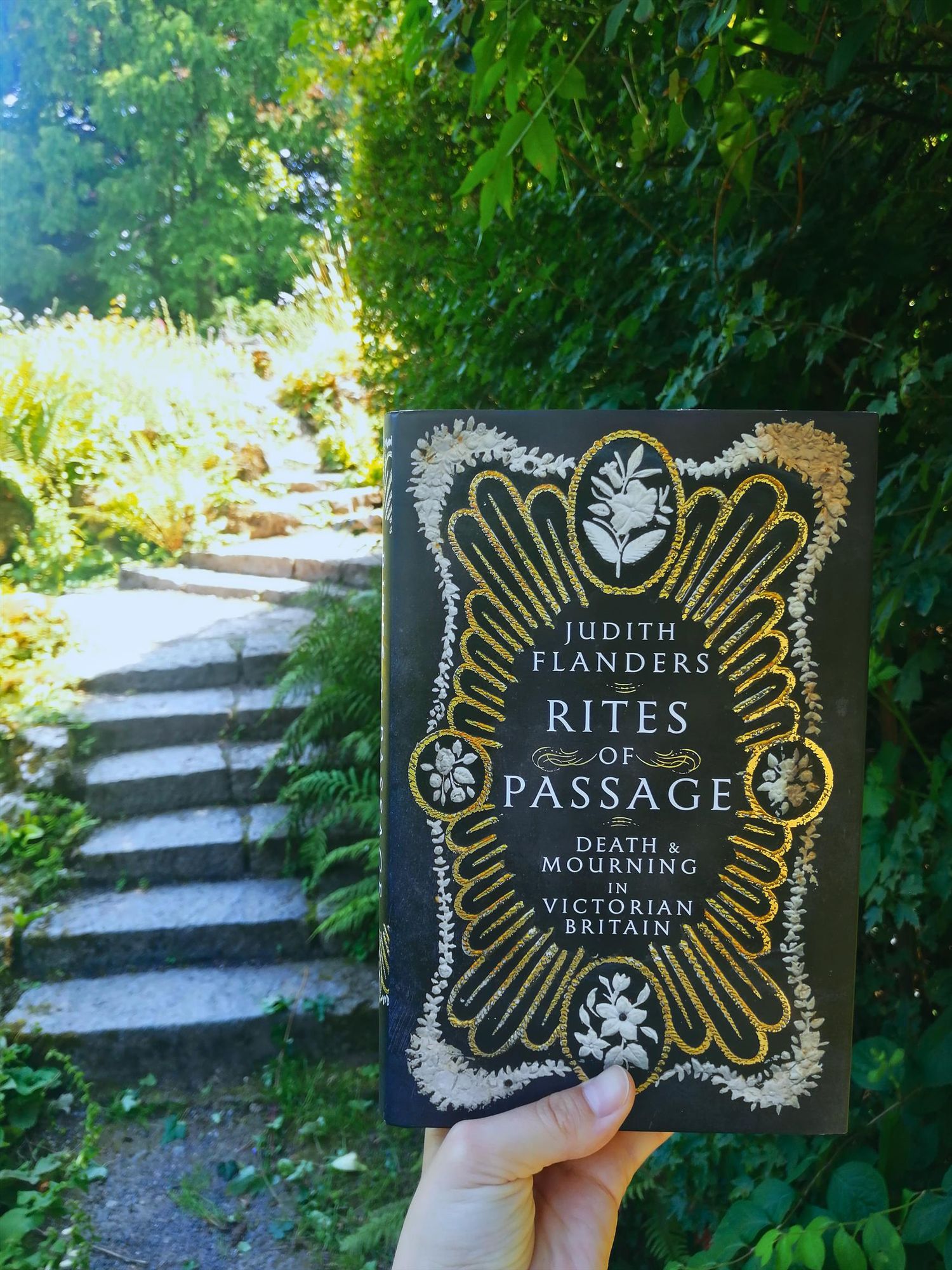 A colour rendition of a photo holding Judith Flanders's book "Rites of Passage: Death & Mourning in Victorian Britain" in the Botanical Gardens, the background shows stone stairs next to a dark and looming wall of ivy (same photo as the left, but in colour)