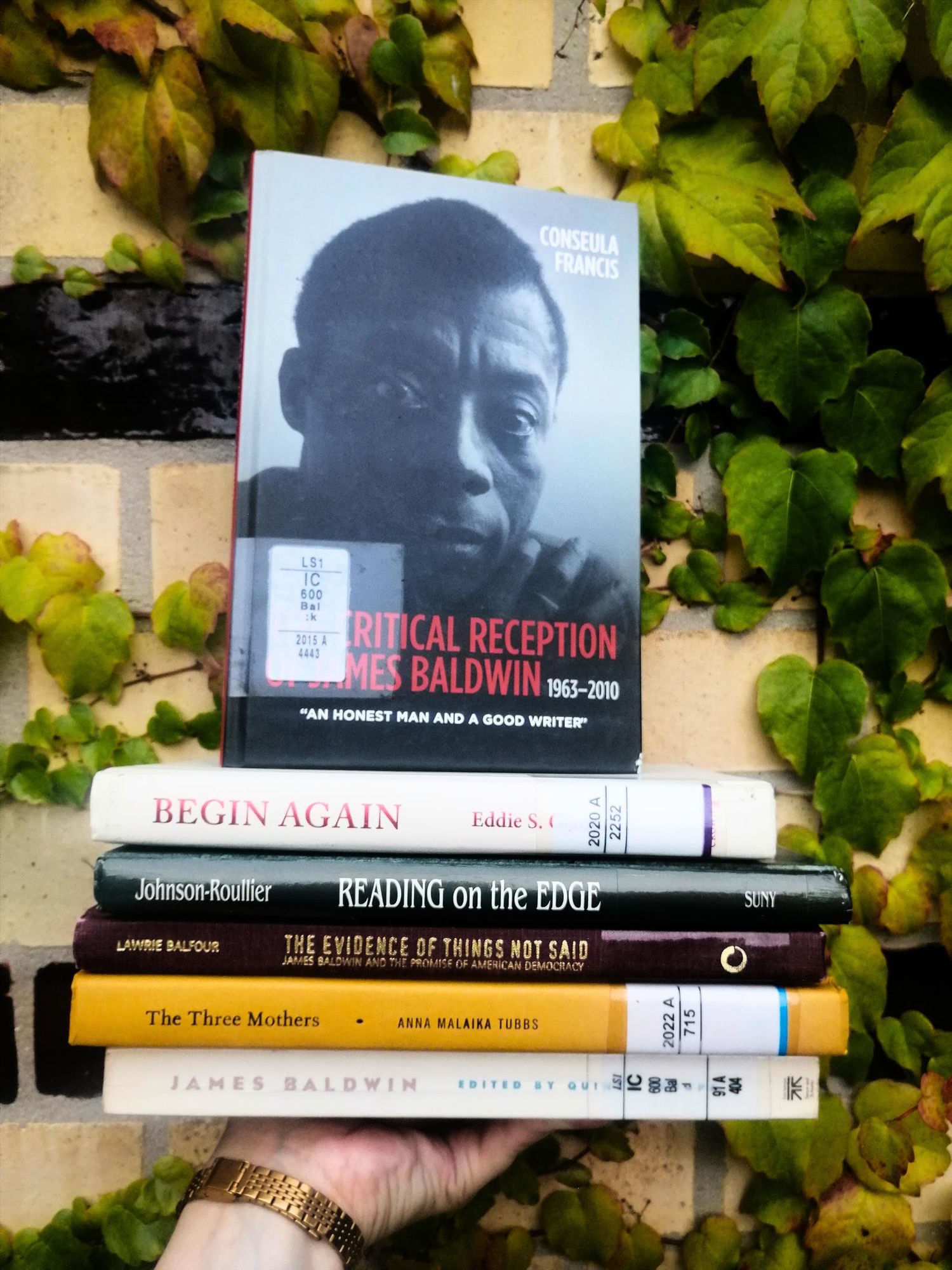 Holding a stack of books on Baldwin with the book "Critical Reception of James Baldwin, 1963-2010" by Consuela Francis on top, which shows a photograph of Baldwin. The background is a yellow-bricked wall overgrown with ivy